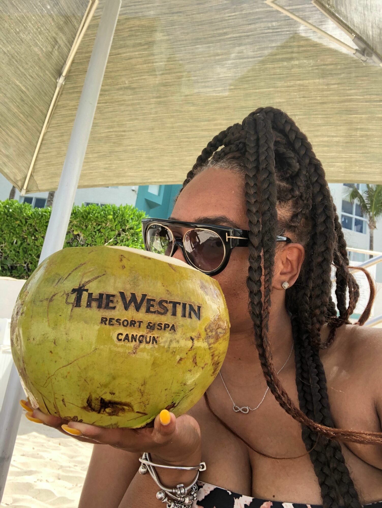 Person with braided hair and sunglasses holding a yellow coconut drink with a straw outdoors.
