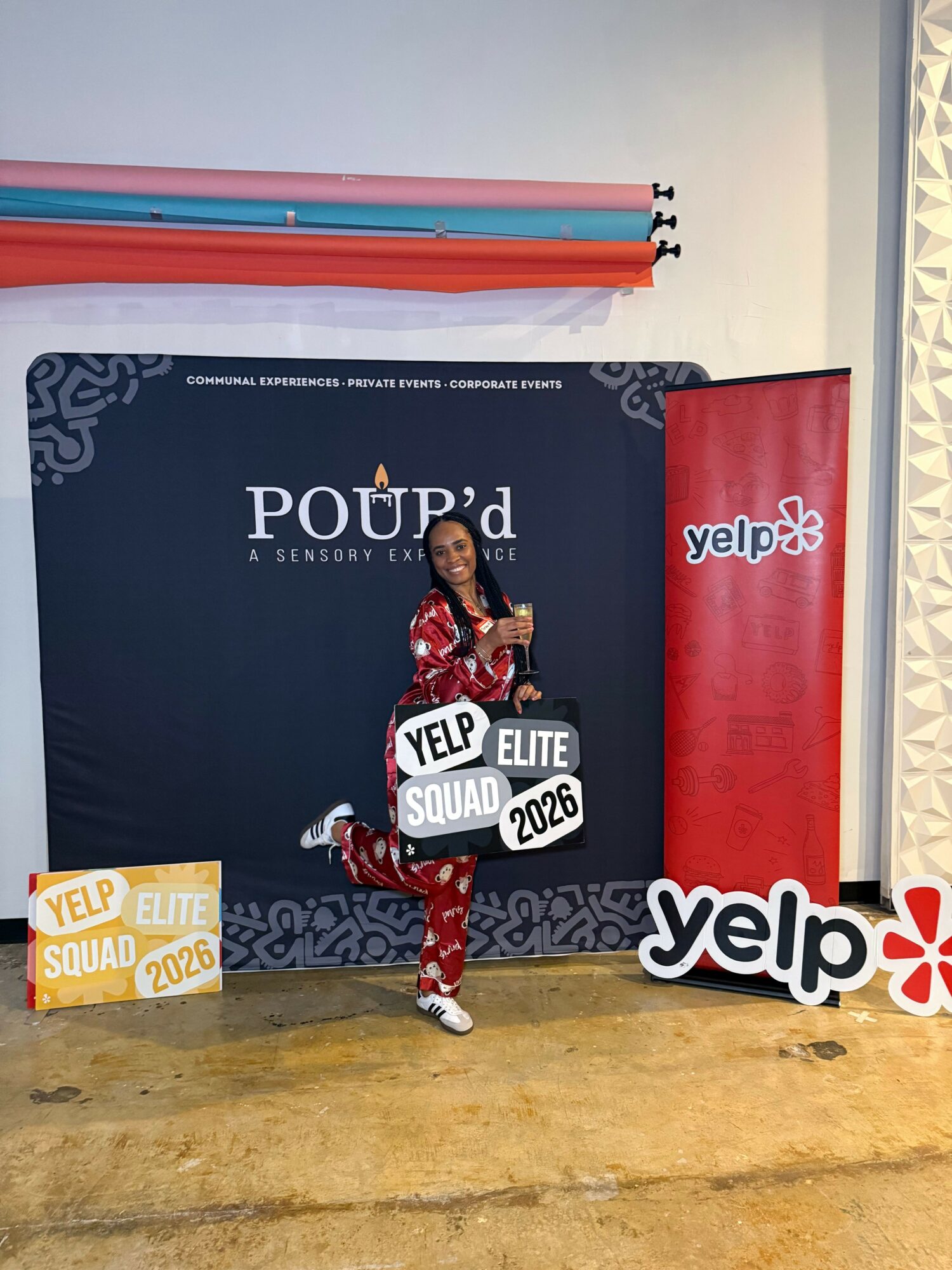 A person in a red and black patterned outfit standing in front of a Yelp event display with banners and signs, on a yellowish ground.
