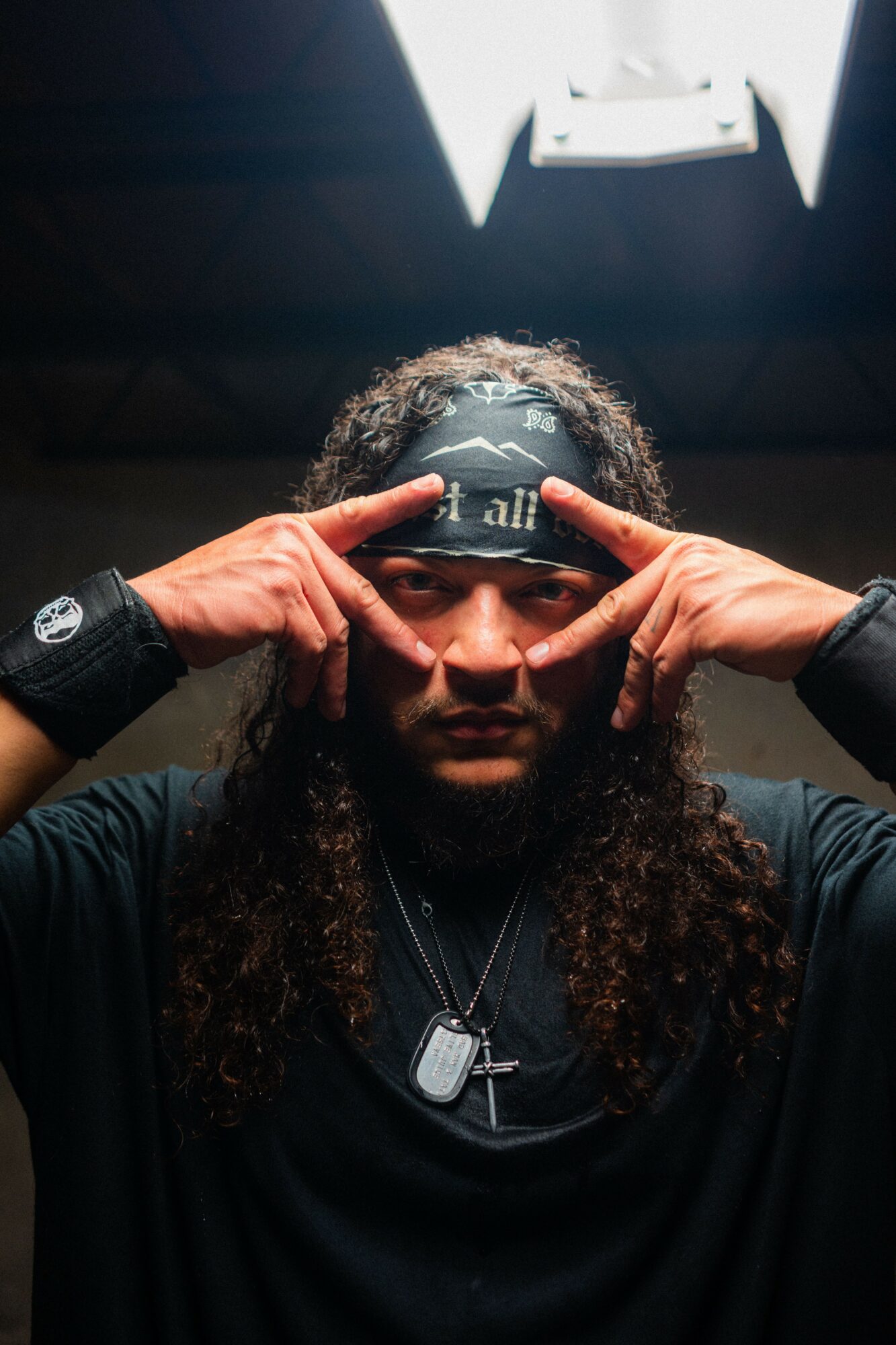 Person with long curly hair wearing a headband, making a gesture with hands near face, dark background, bright light above