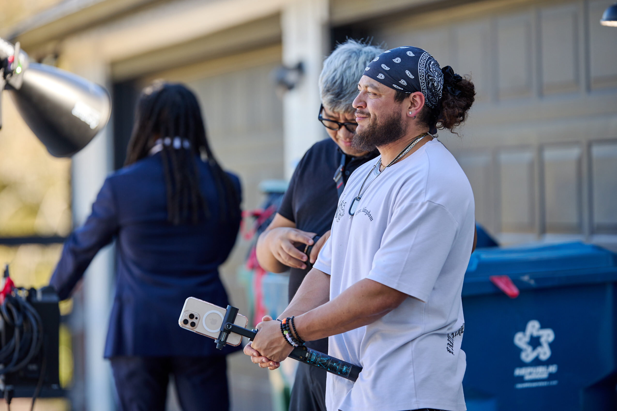 Three people standing outdoors, one with a bandana, holding a tool, and two others engaged in conversation, with a garage door in the background.