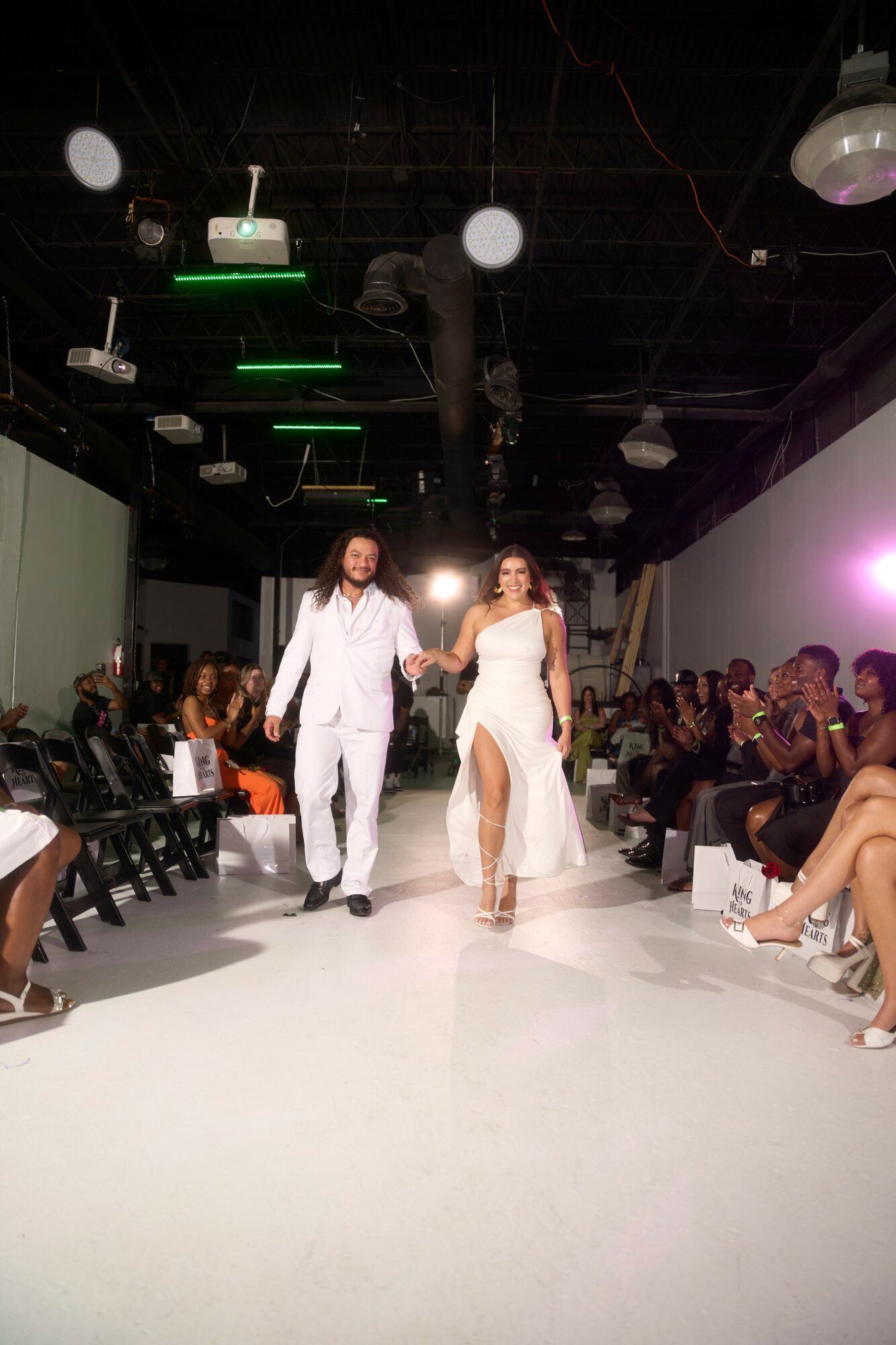 Two models walk down a runway in white outfits, audience seated on both sides, dark ceiling with lights and equipment