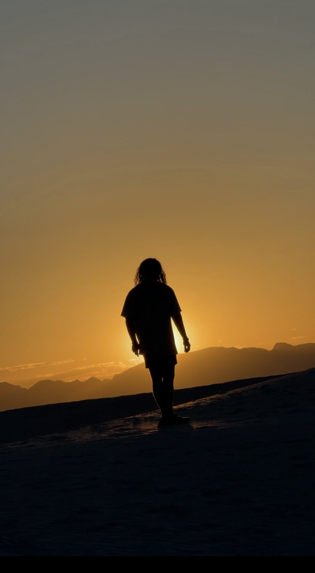 Person walking on a hill during sunset with a colorful sky and distant mountains.