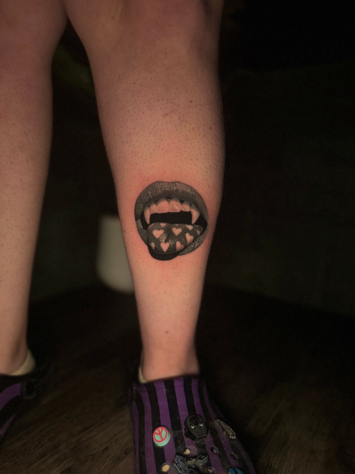 Tattoo of an open mouth with sharp teeth on person's lower leg, wearing purple and black striped sock, dark background.