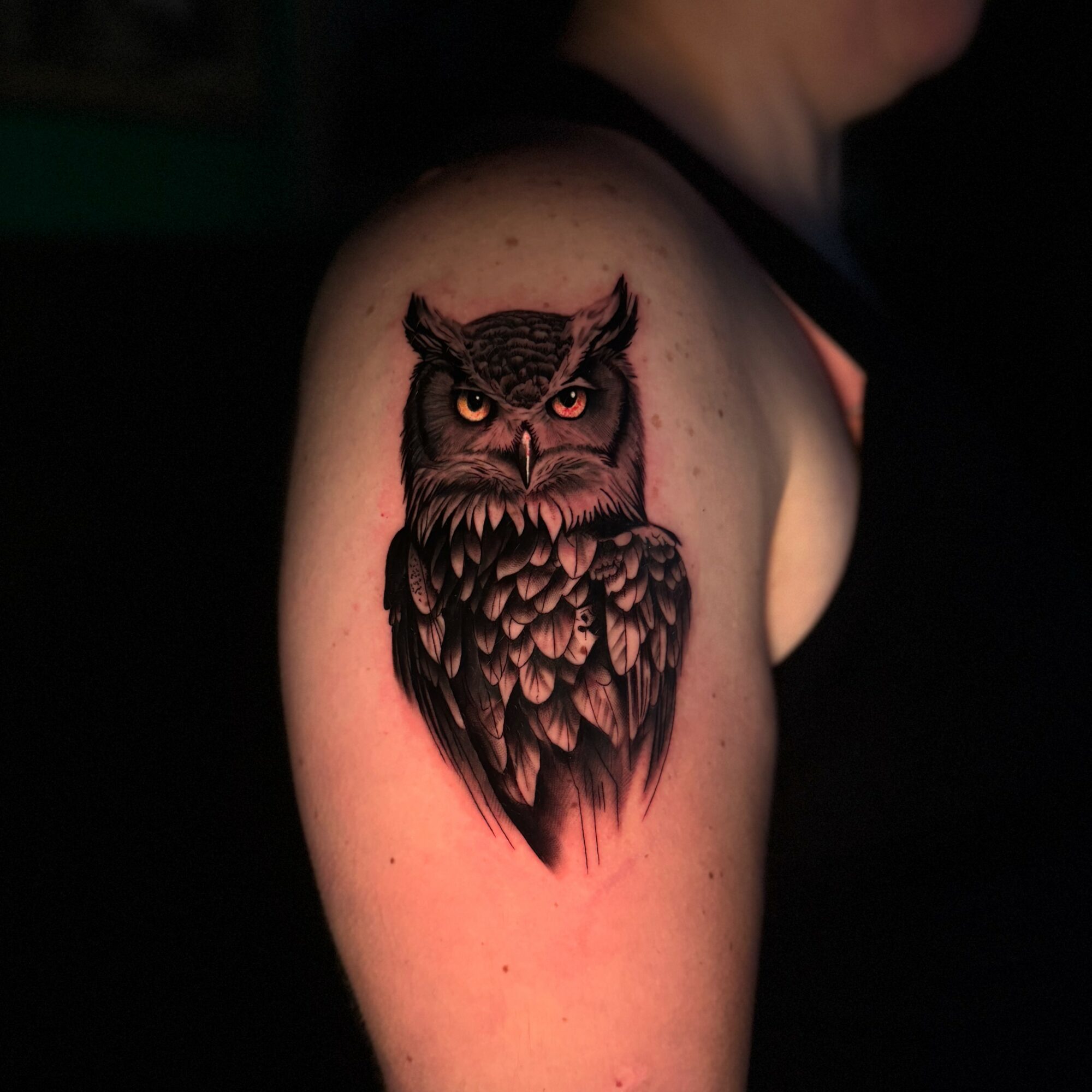 Tattoo of an owl with detailed feathers on person's upper arm, facing forward with intense eyes.