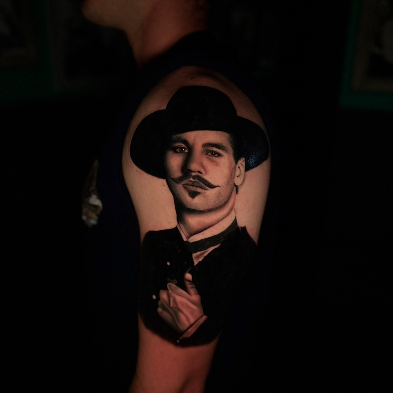 Tattoo of a man with a mustache, wearing a hat, holding a cigarette, on person's arm.