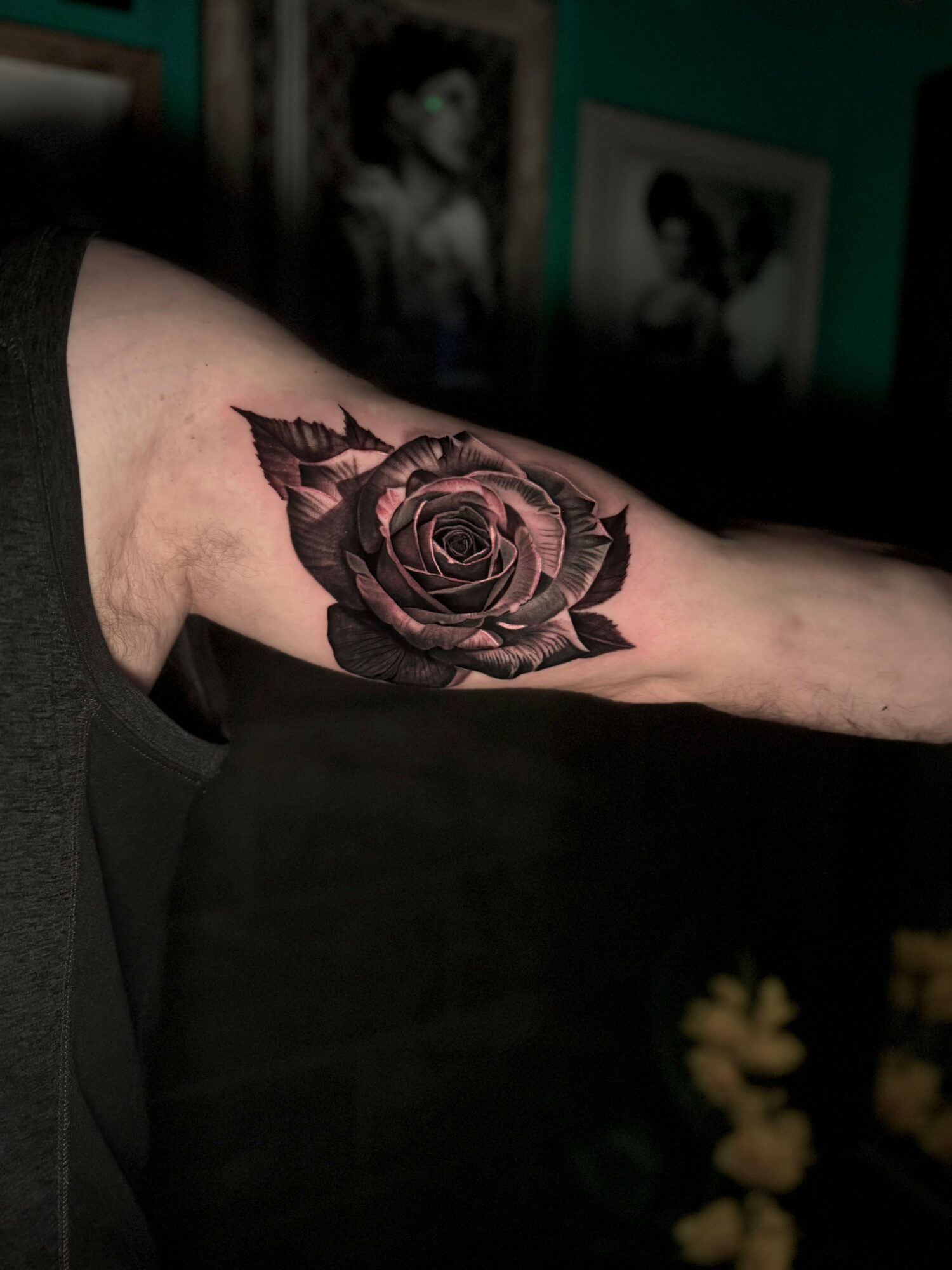 Black and gray rose tattoo on person's upper arm, with framed pictures in background.