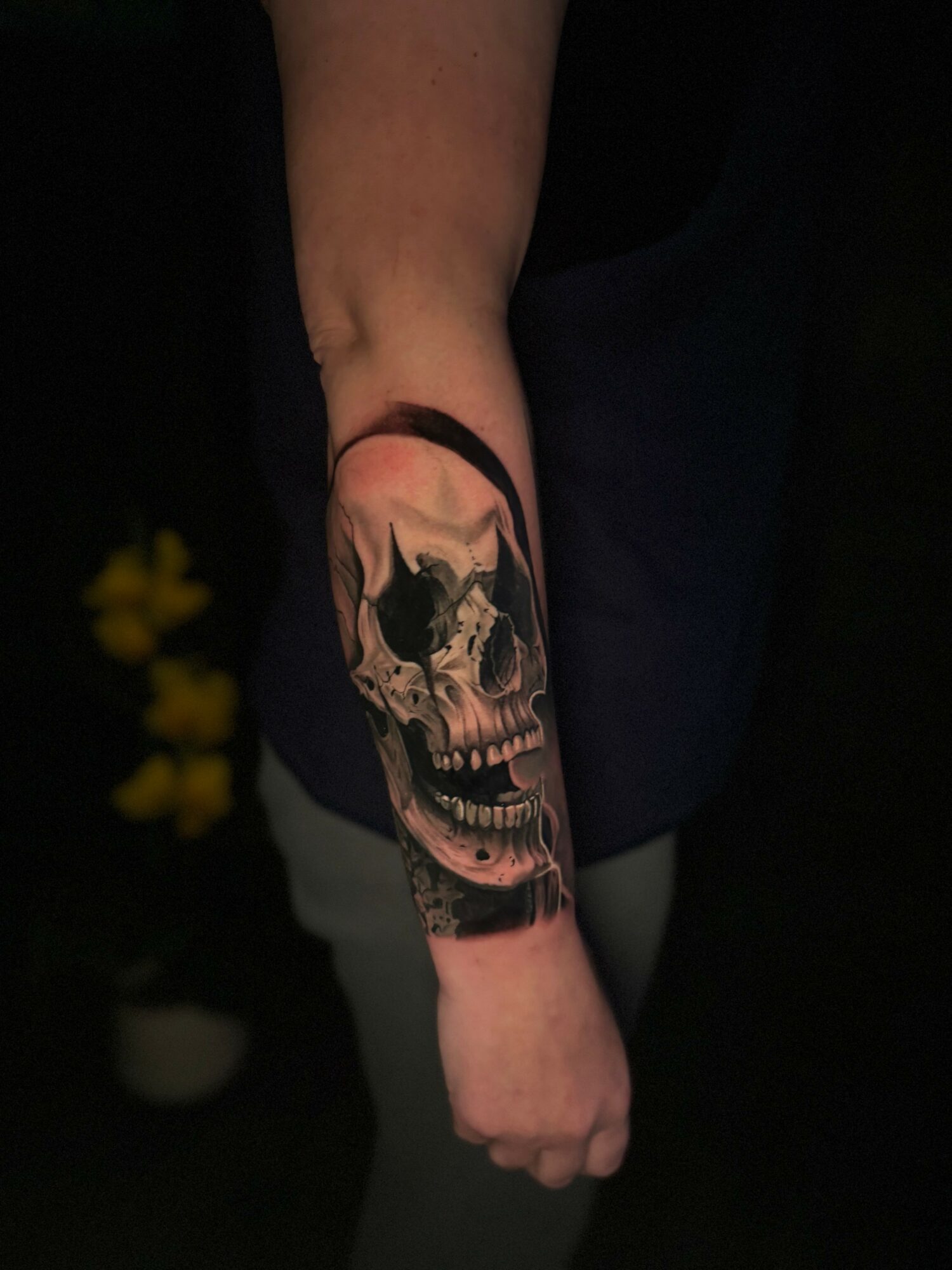 Person's arm with a detailed skull tattoo, dark background, and yellow flowers partially visible.