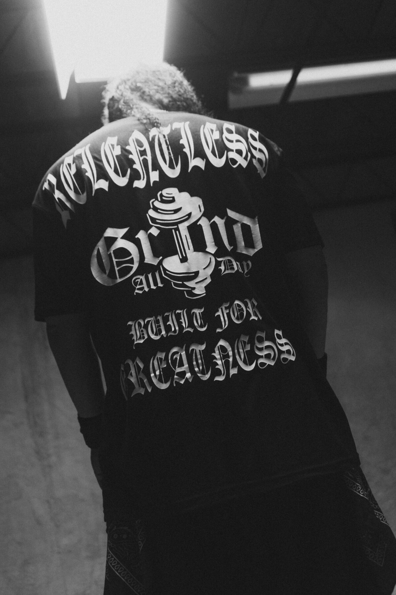 Person with curly hair wearing a black shirt with bold white text and graphic design on the back, indoors with light source above.
