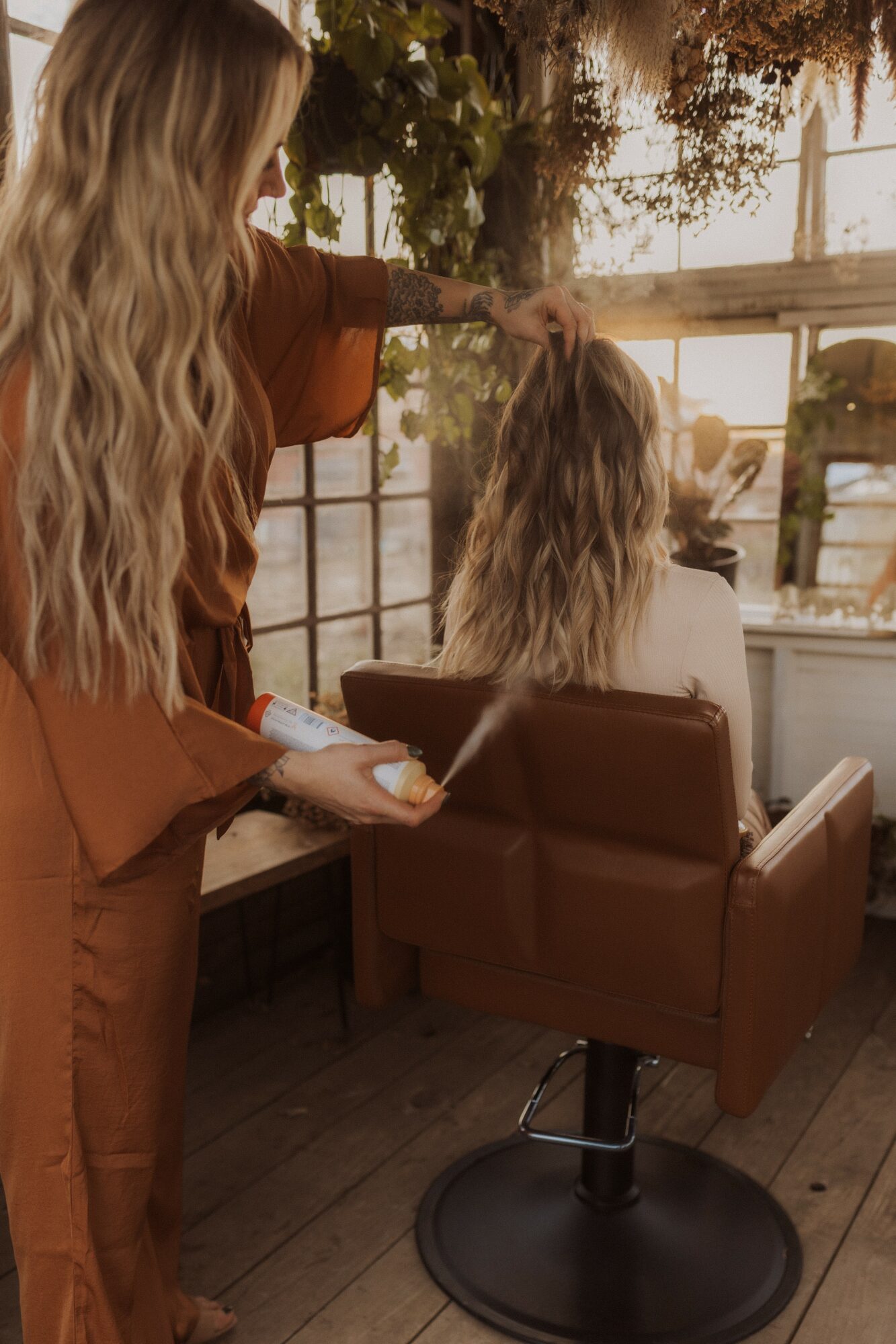 Hairdresser styles a woman's long, wavy hair in a sunlit salon with large windows and plants.