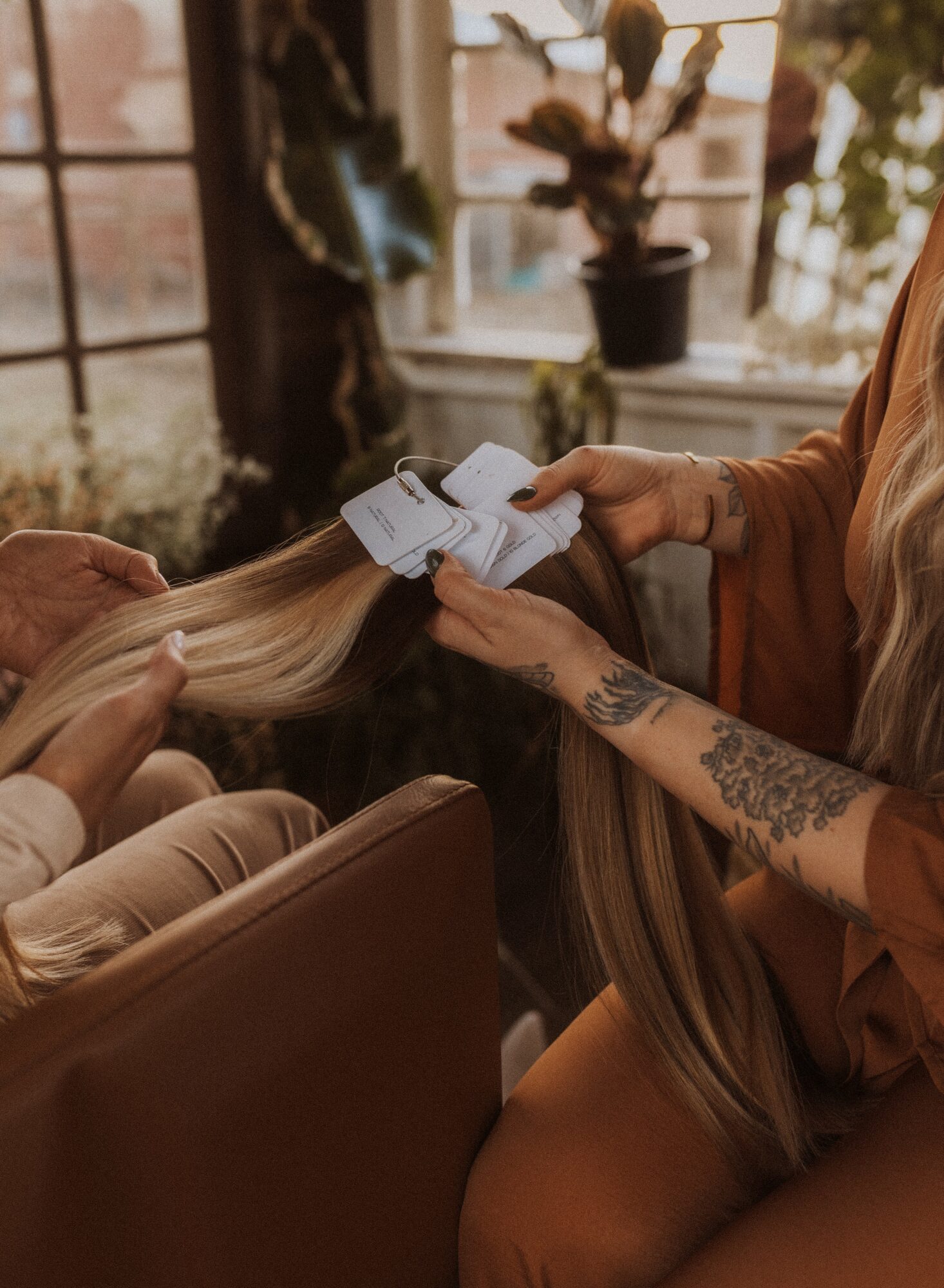 Person with tattooed arm holding a hair extension tool, working on long blonde hair in a cozy room with plants and large windows.