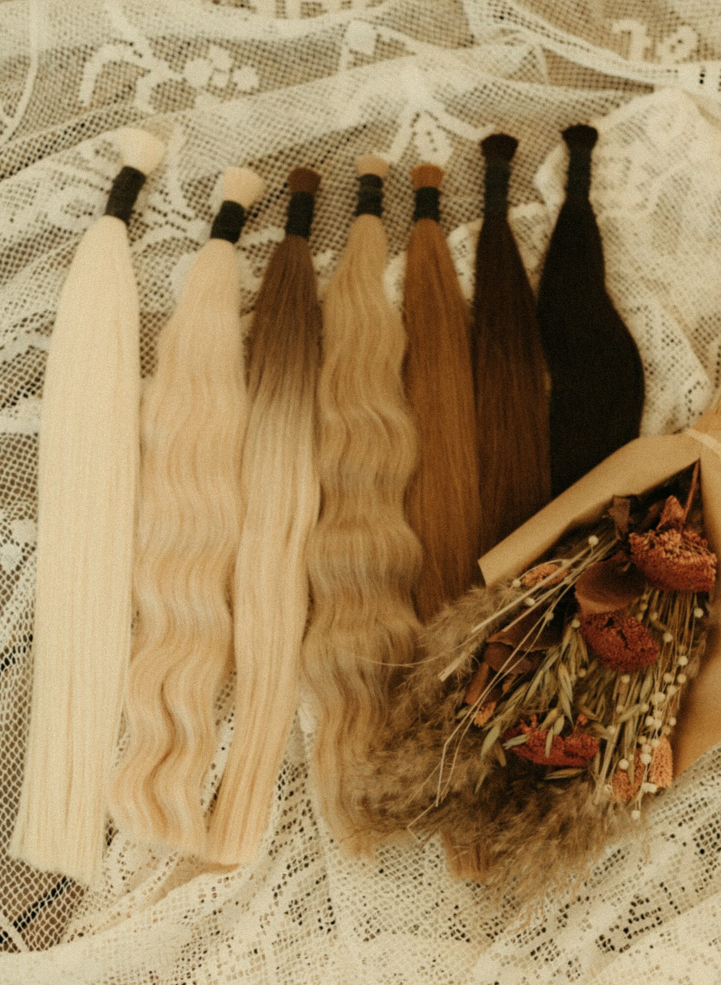 Six bundles of hair extensions in various shades, arranged on lace fabric with a small bouquet of dried flowers and a rolled paper.
