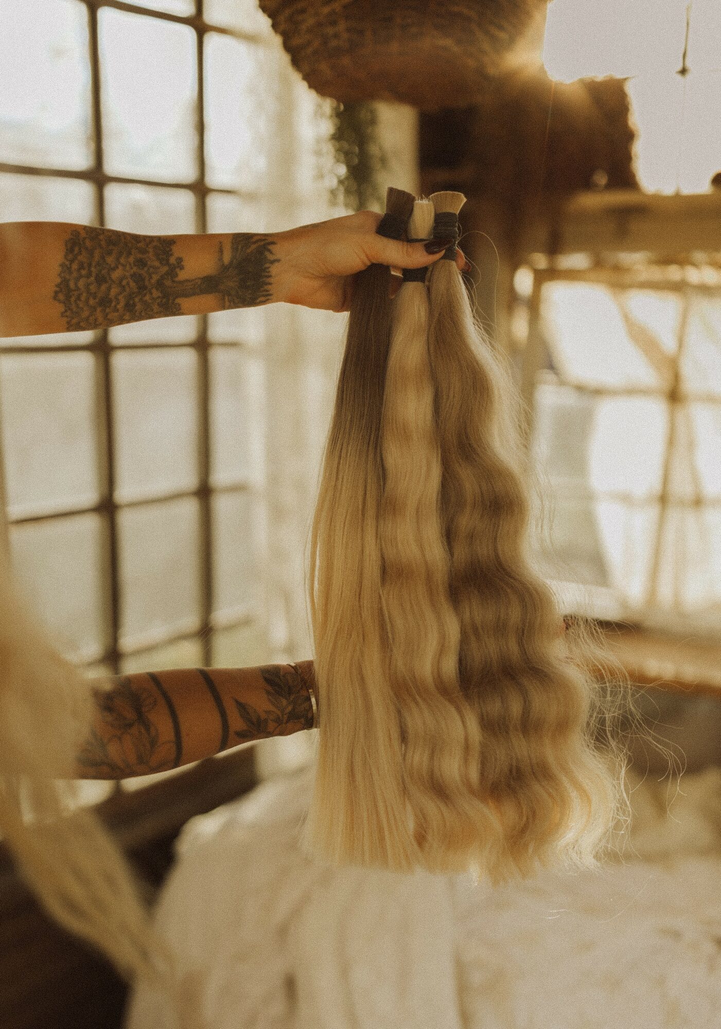 Person holding long, wavy blonde hair extension in a room with large windows and warm lighting.