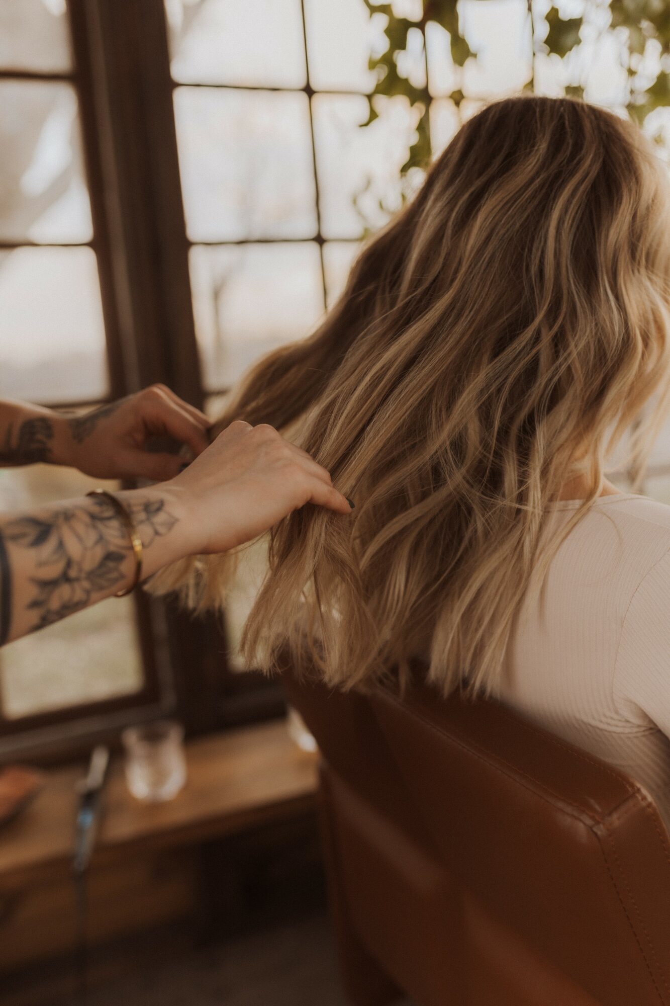 Person with long, wavy blonde hair sitting in a chair, with a stylist working on their hair, in a room with large windows.