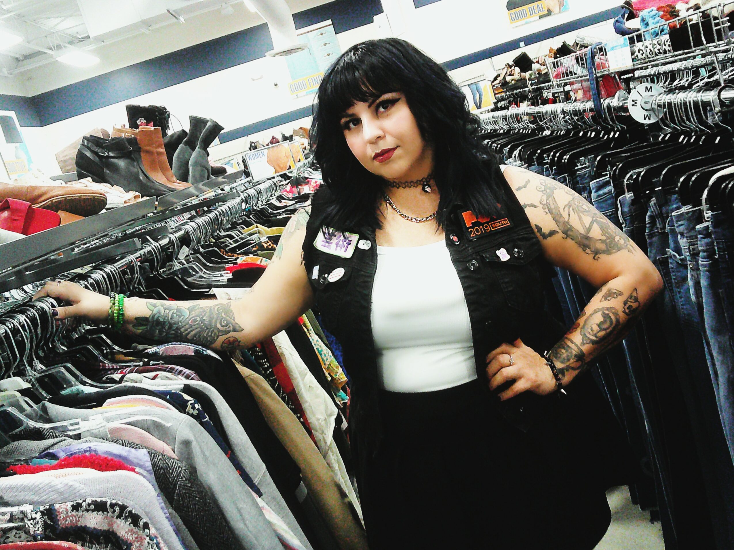 Woman with black hair and tattoos shopping for clothes in a store, surrounded by racks of clothing.
