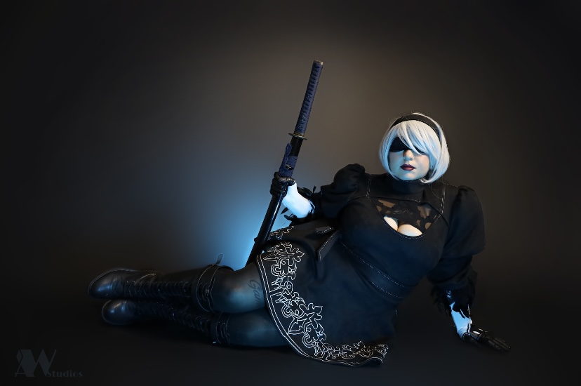 Person with short blonde hair, black outfit, sitting on the floor holding a sword, against a dark background.