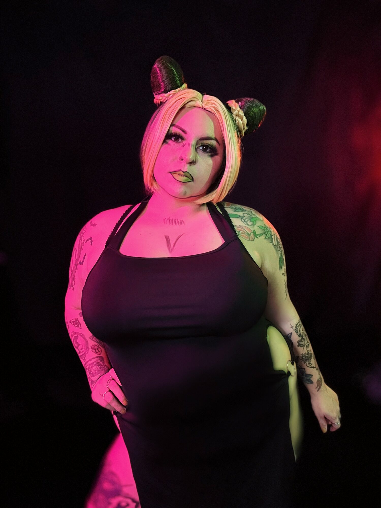 Woman with pink hair, green and black animal ears, tattoos, wearing a black dress, standing in a dark setting with pink lighting.