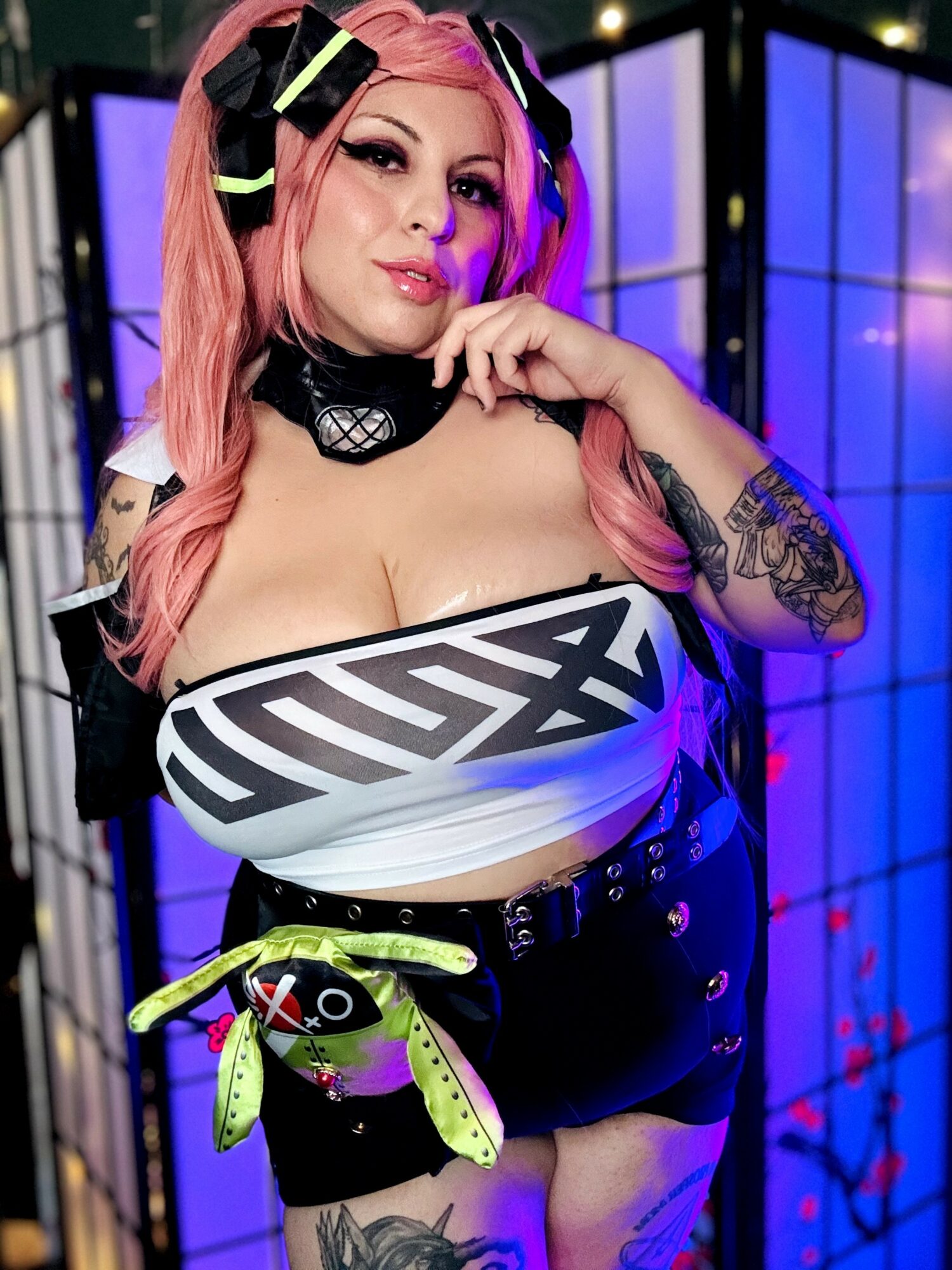 Woman with pink hair and tattoos, wearing a futuristic outfit, in front of colorful illuminated panels.
