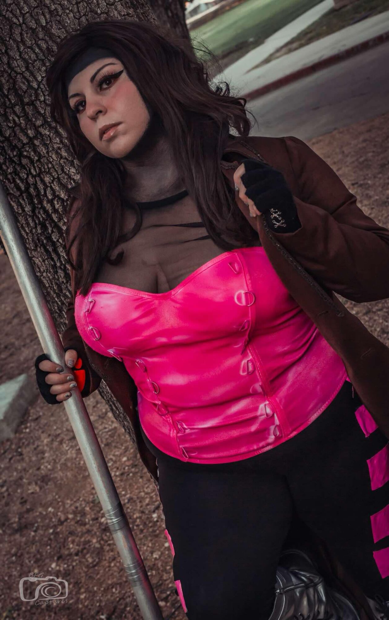 Young woman with dark hair and makeup, wearing a pink top, black pants, and a brown jacket, standing outdoors near a tree.