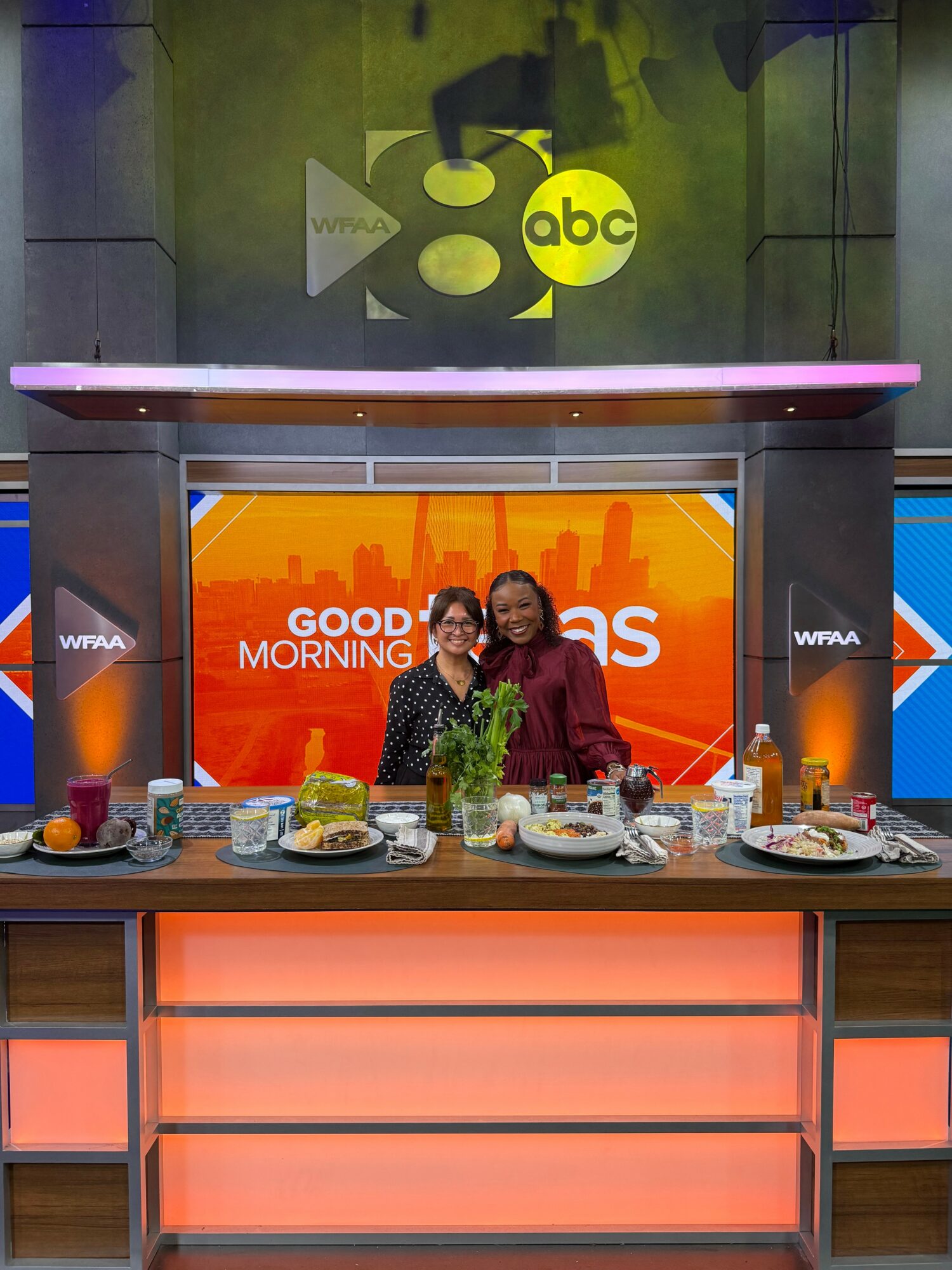 Two women stand behind a breakfast buffet on a TV set with a cityscape background and a bright orange screen reading 'Good Morning'.