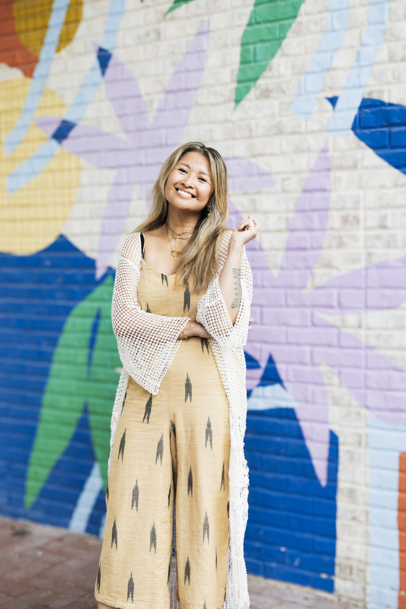 Young woman with long hair smiling in front of a colorful mural wall, wearing a yellow dress and a white cardigan.