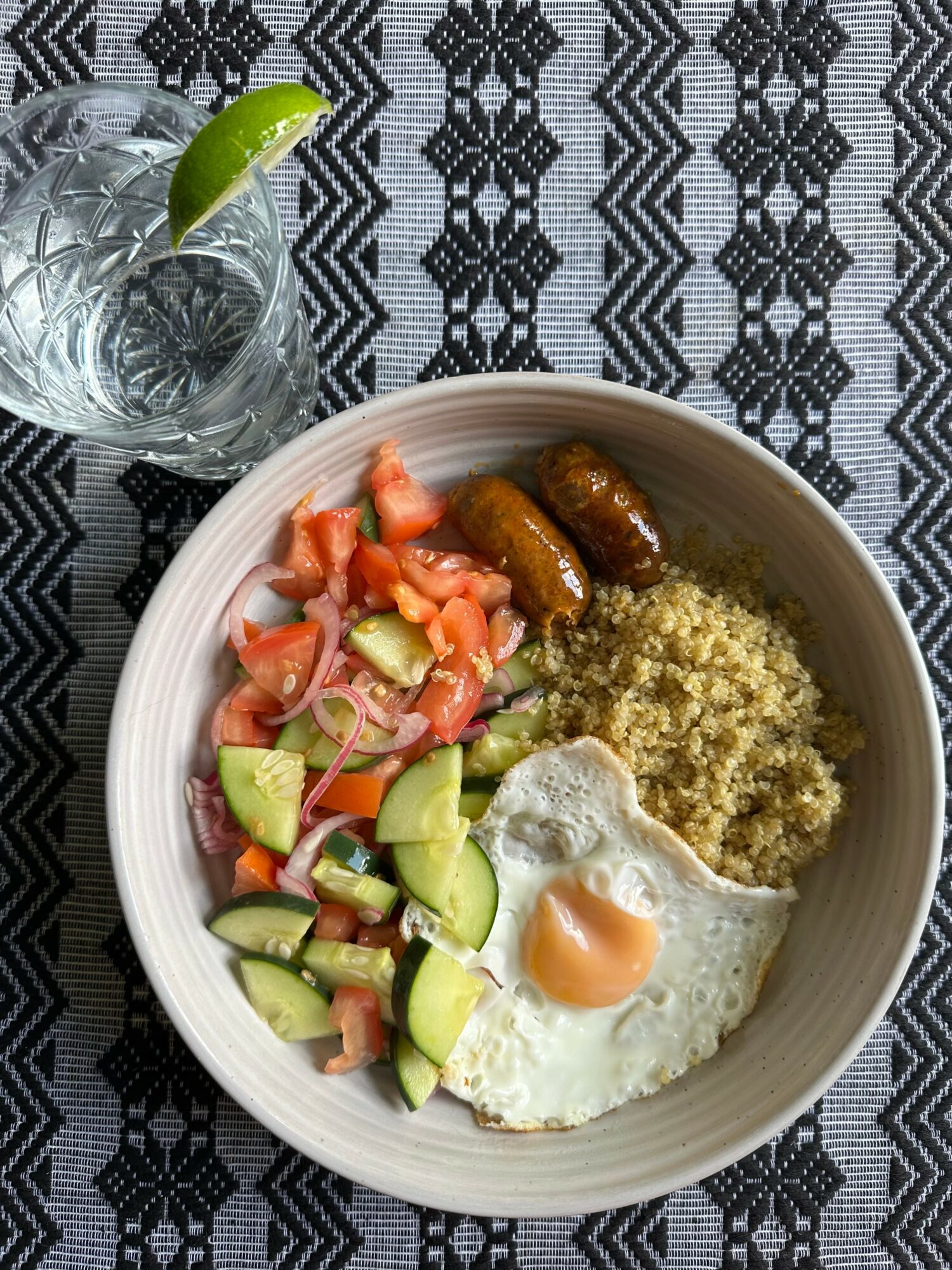 Bowl with fried egg, couscous, cucumber, tomato salad, and two small sausages on a patterned tablecloth, with a glass of water and lime wedge nearby.