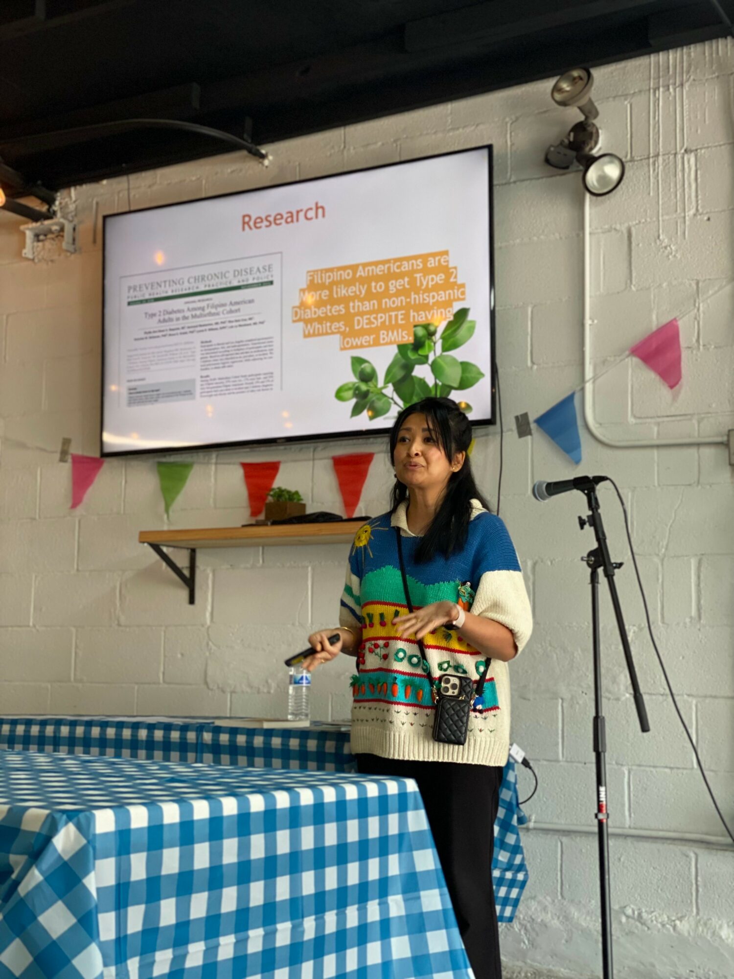 Young woman standing in front of a presentation screen, holding a water bottle, with colorful bunting and a table with a blue checkered tablecloth nearby.