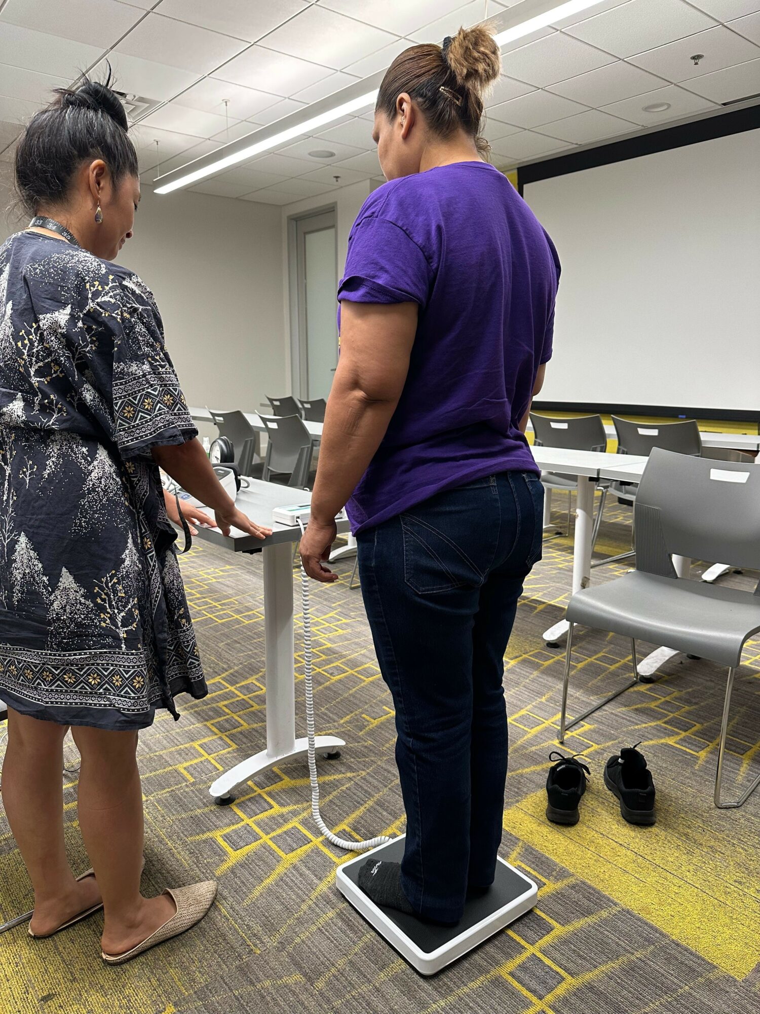 Person standing on a scale in a room with chairs and tables, with another person sitting nearby, in an office or classroom.