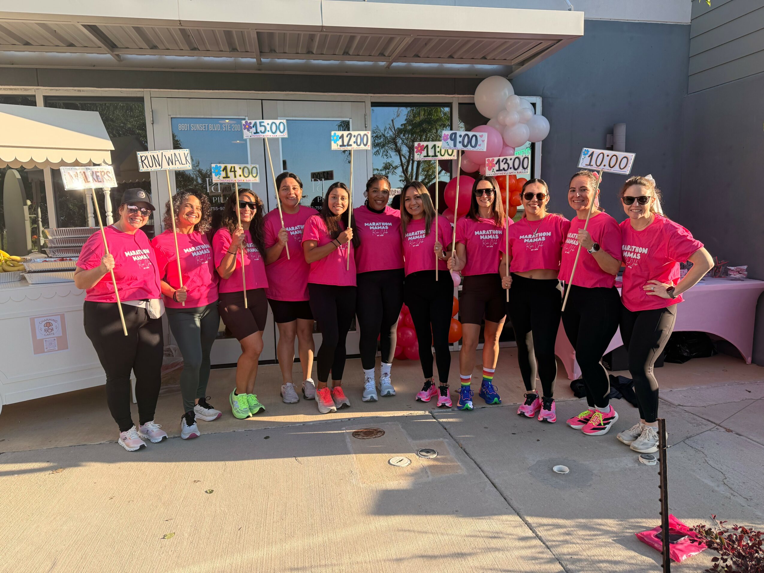 Group of people in pink shirts holding signs with times, standing outdoors in front of a building, smiling.
