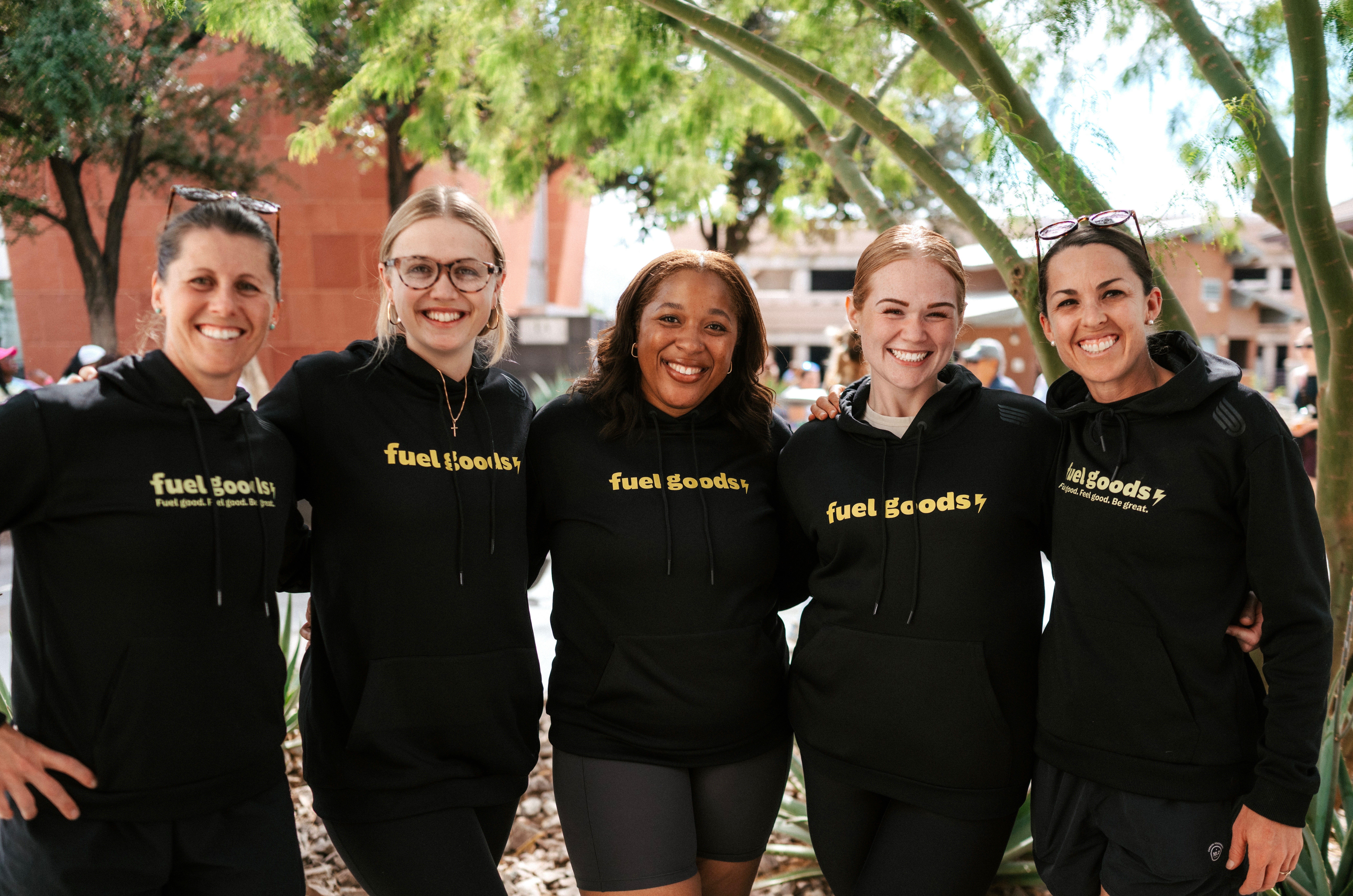 Five women standing outdoors under trees, smiling, wearing matching black hoodies with yellow text, in a park-like setting.