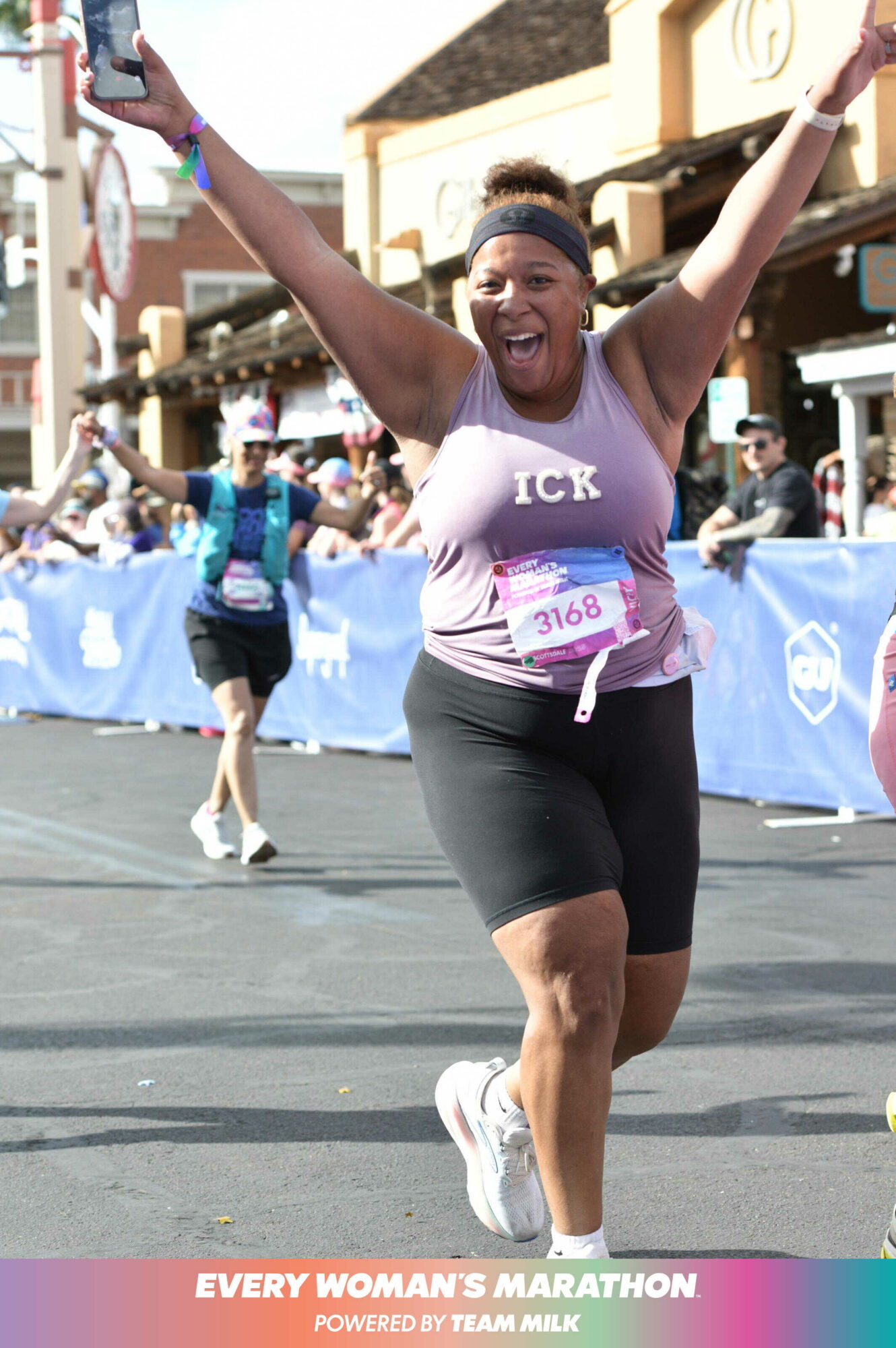 Woman celebrating with arms raised during marathon race on city street, smiling, wearing a pink tank top and black shorts.