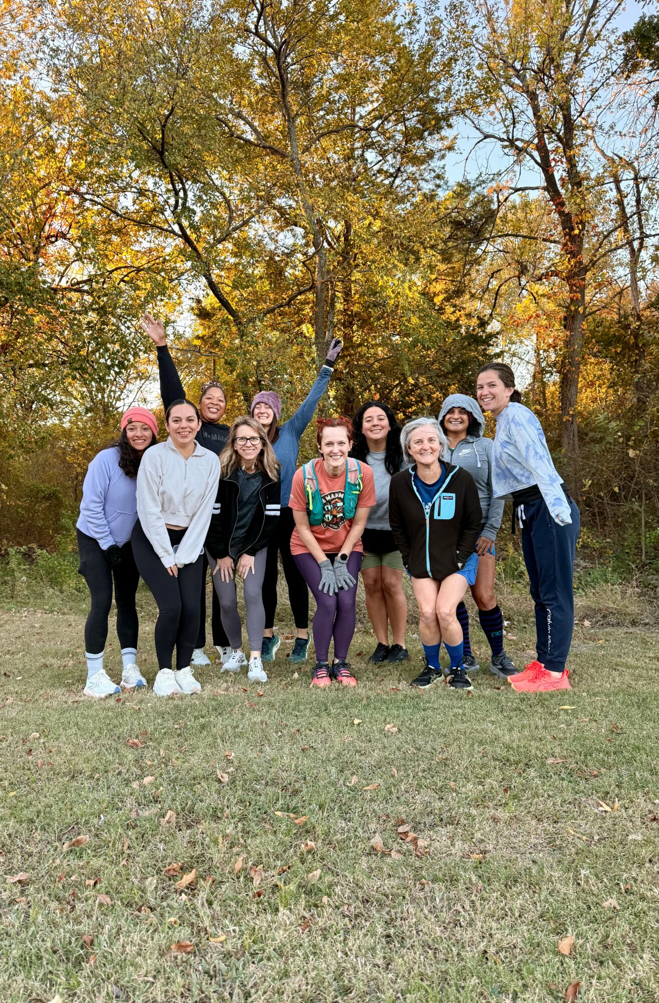 Group of ten people standing outdoors on grass with trees and autumn foliage in background.