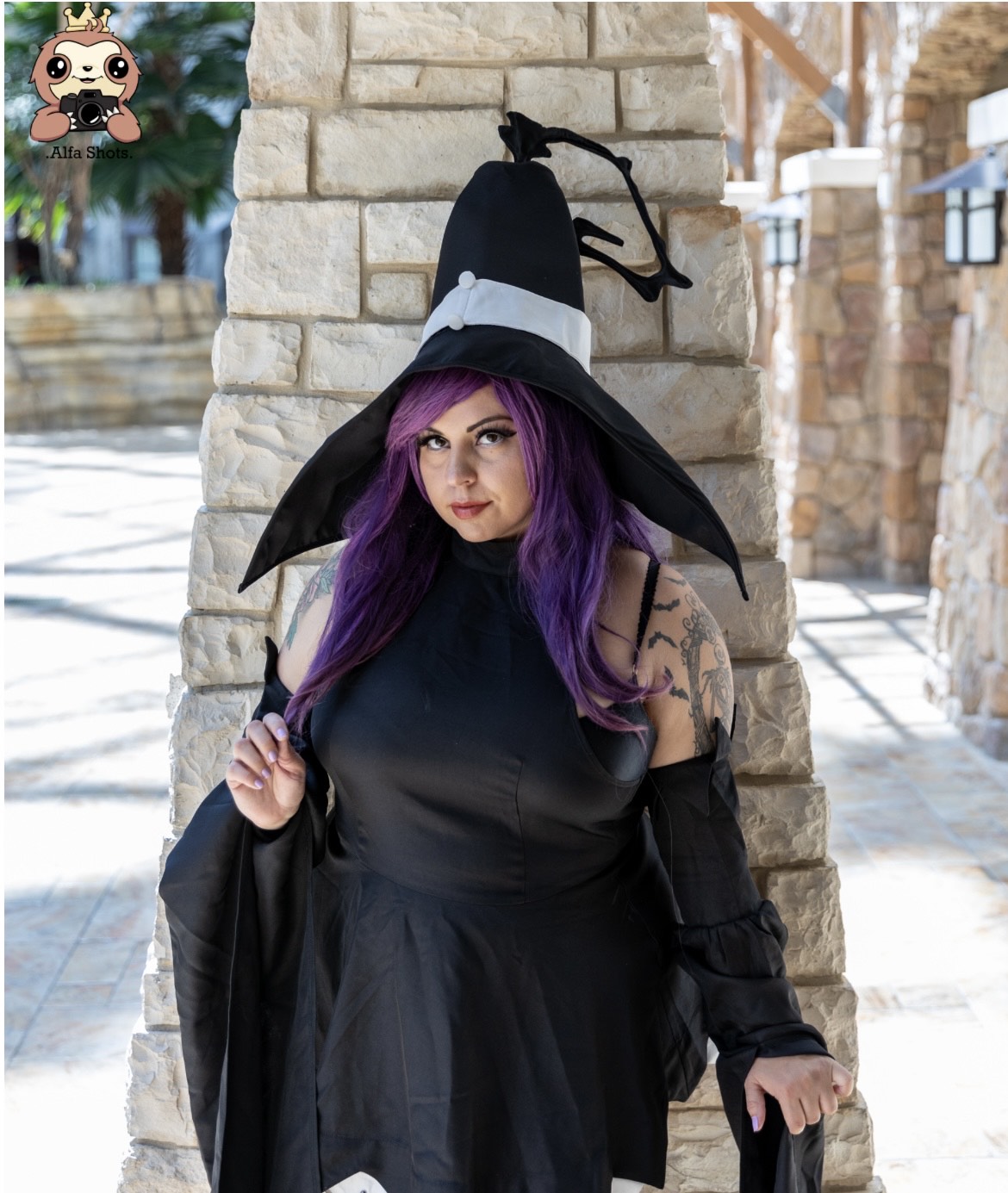 Woman with purple hair in a witch costume, wearing a black hat, standing against a stone wall outdoors.