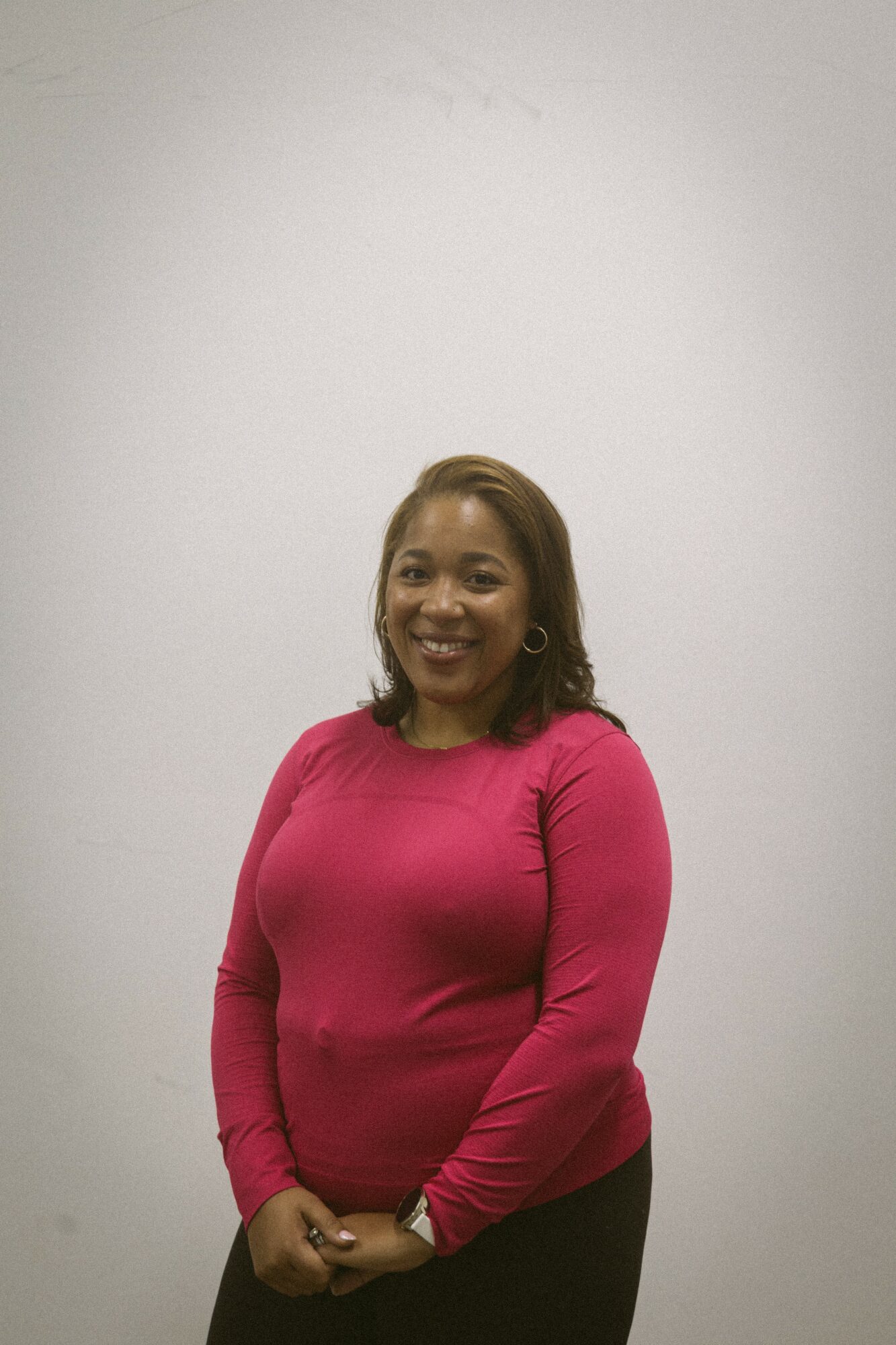 Smiling woman with shoulder-length hair wearing a pink long-sleeve top, standing against a plain white background.