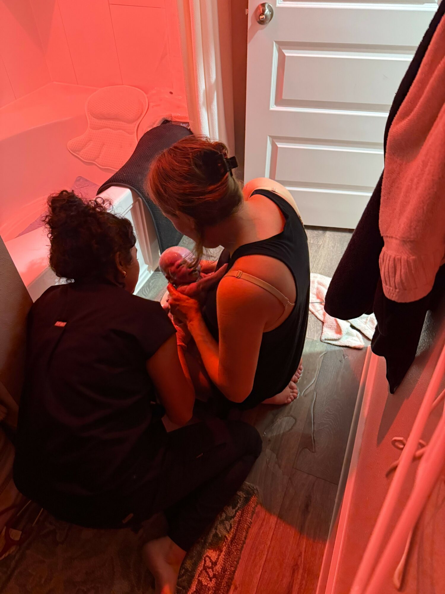 Two people sitting on the floor near a door, one with long hair and the other with curly hair, in a room with a wooden floor and a bed with a pink blanket. The image has a red tint.