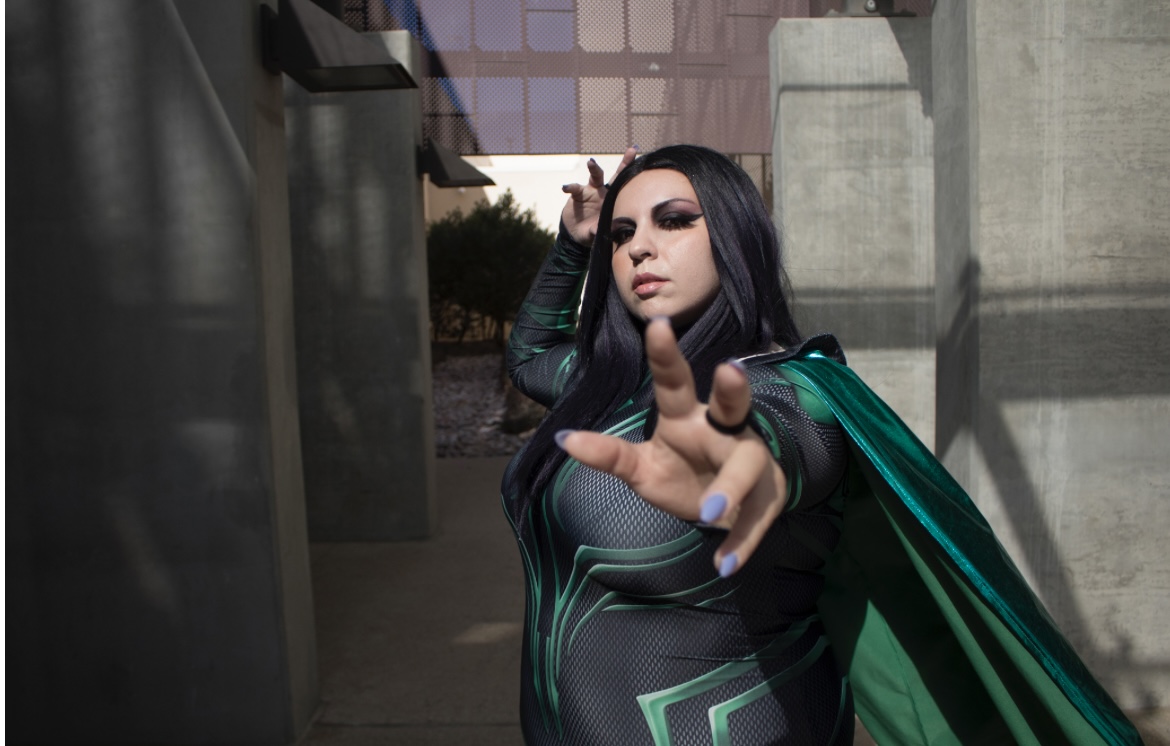 Woman with long dark hair in a futuristic costume, pointing towards the camera outdoors, concrete structures in background.