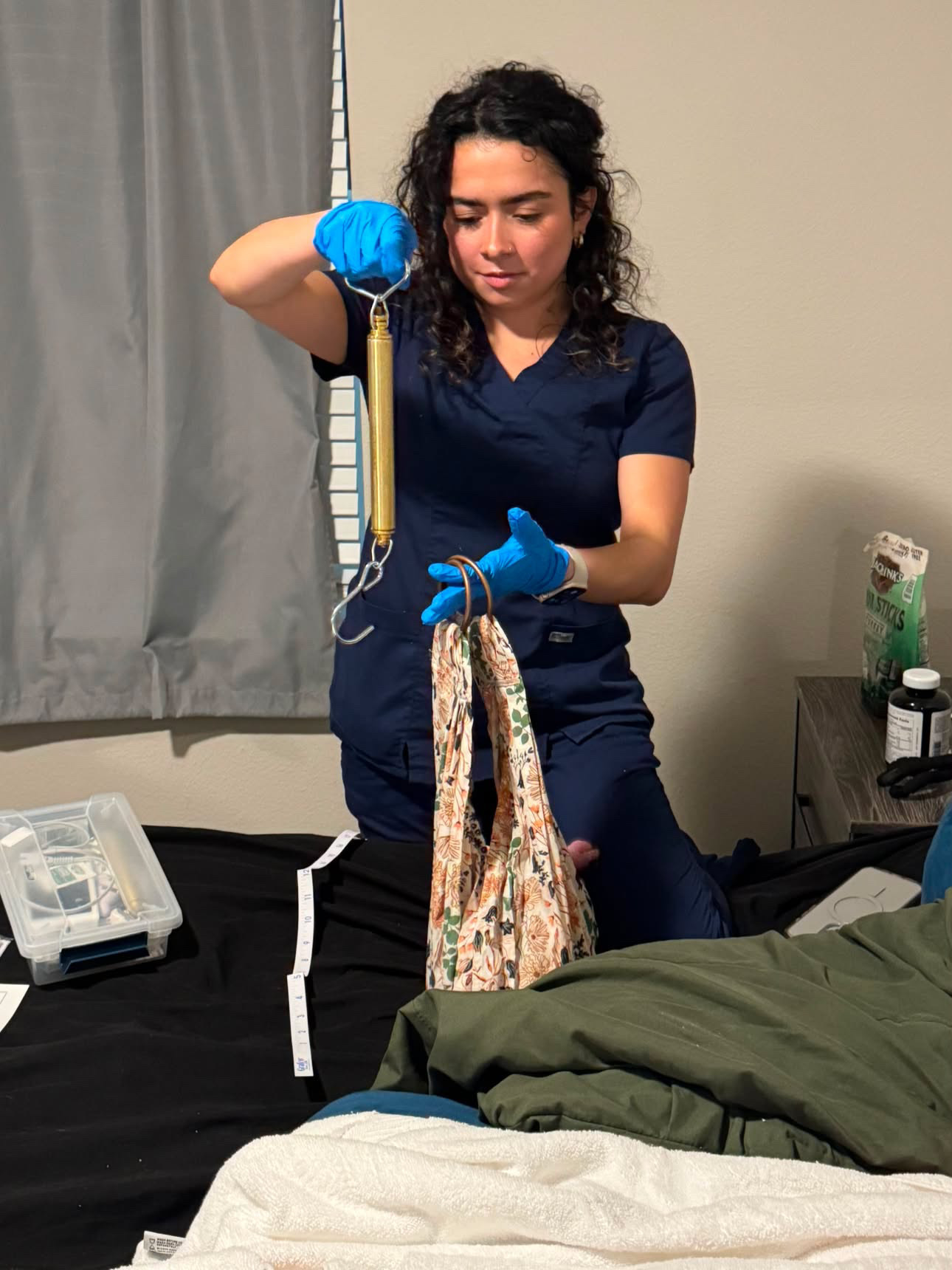 A woman in blue scrubs and gloves holds a medical instrument, standing next to a bed with a bag hanging from her hand.