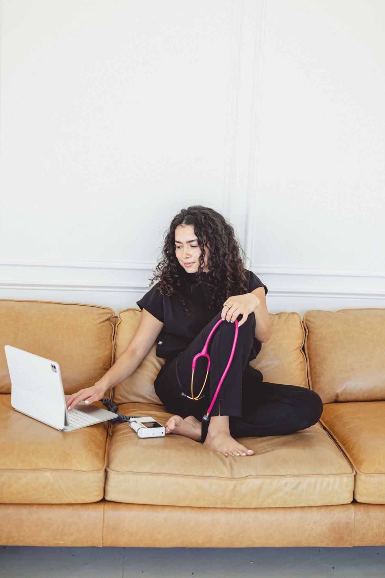 Woman sitting on a sofa with a laptop, holding a pink and black bag, in a room with white walls.