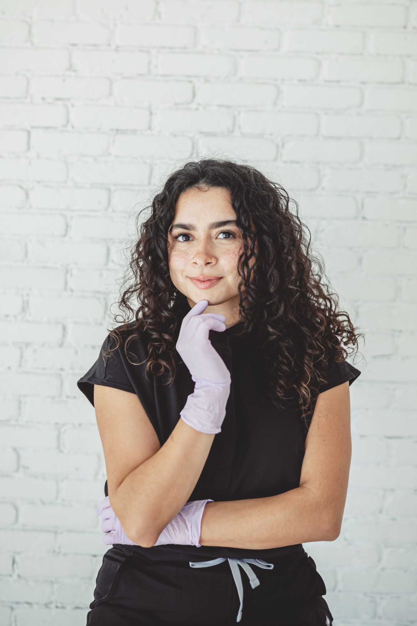 Young woman with curly hair wearing a black shirt and light purple gloves, standing against a white brick wall.