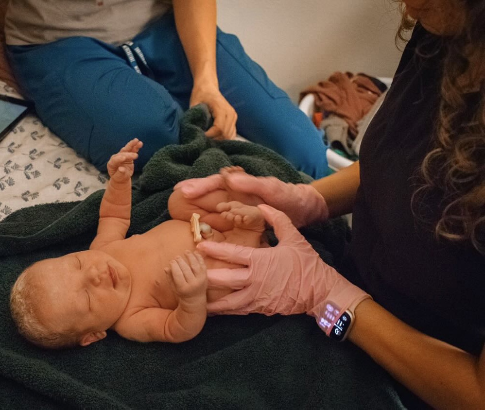 A newborn baby lies on a blanket, being examined by a healthcare professional wearing pink gloves, with a person sitting nearby.