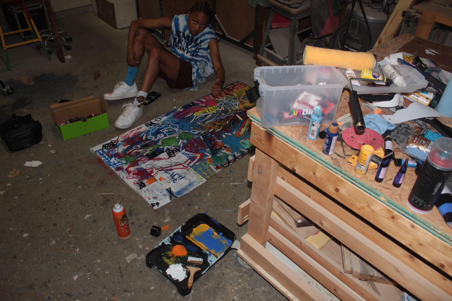Person sitting on the floor with legs extended, surrounded by art supplies and a colorful canvas, in a cluttered workspace.