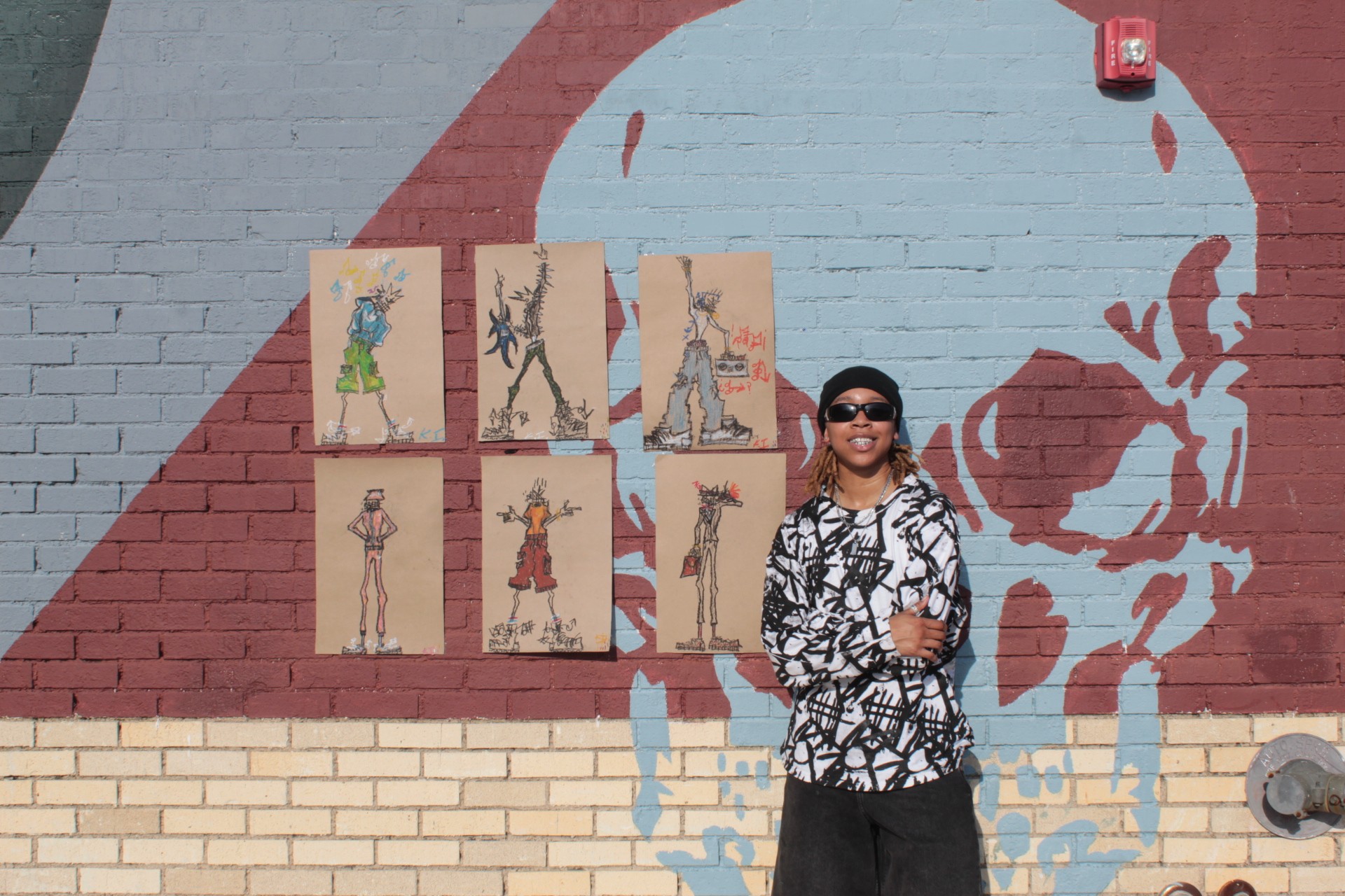 Person wearing sunglasses and patterned shirt standing in front of a mural with a skull and six paintings on a brick wall.