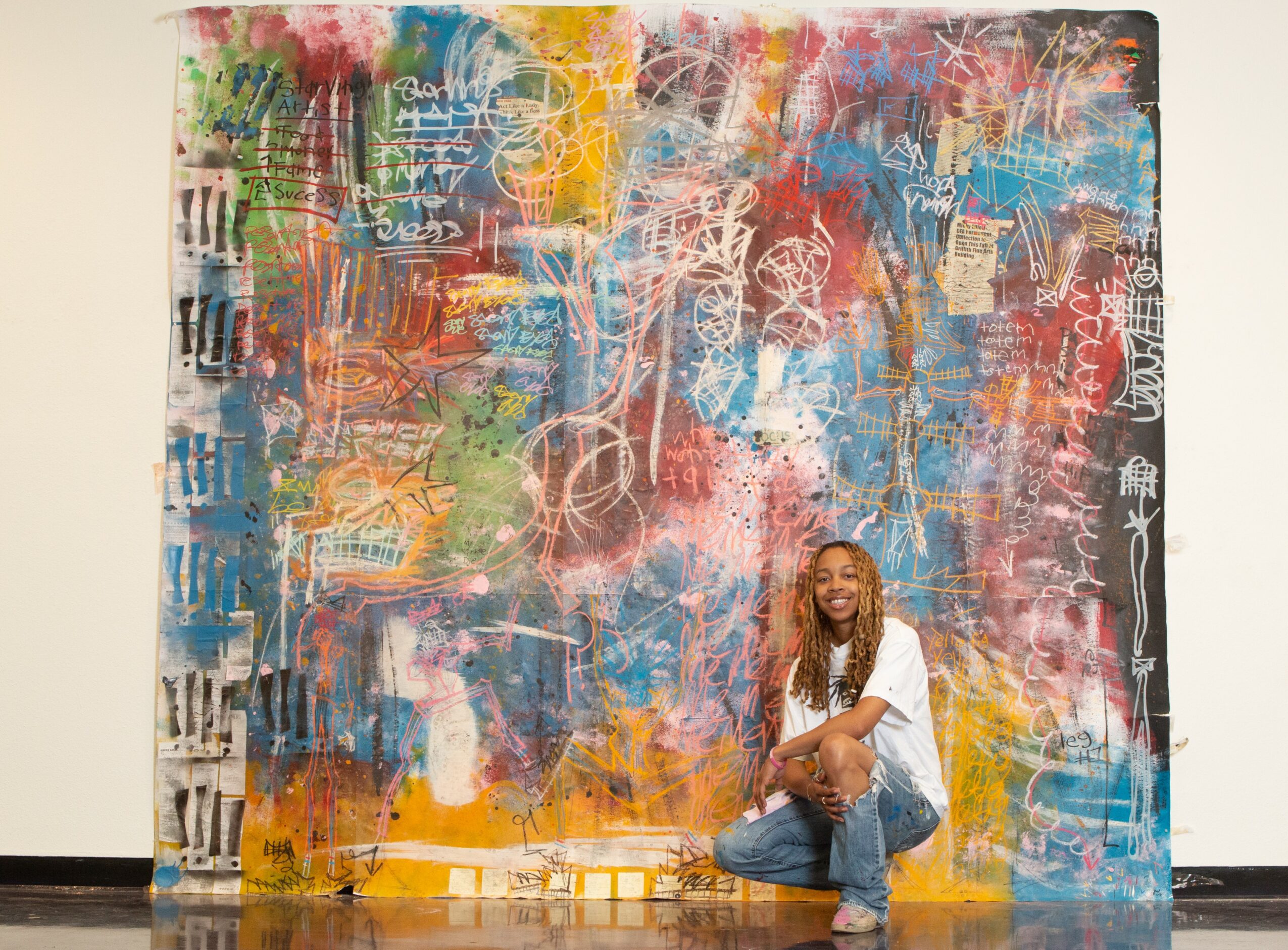 Person sitting on the floor in front of a large colorful abstract painting with various shapes and textures.