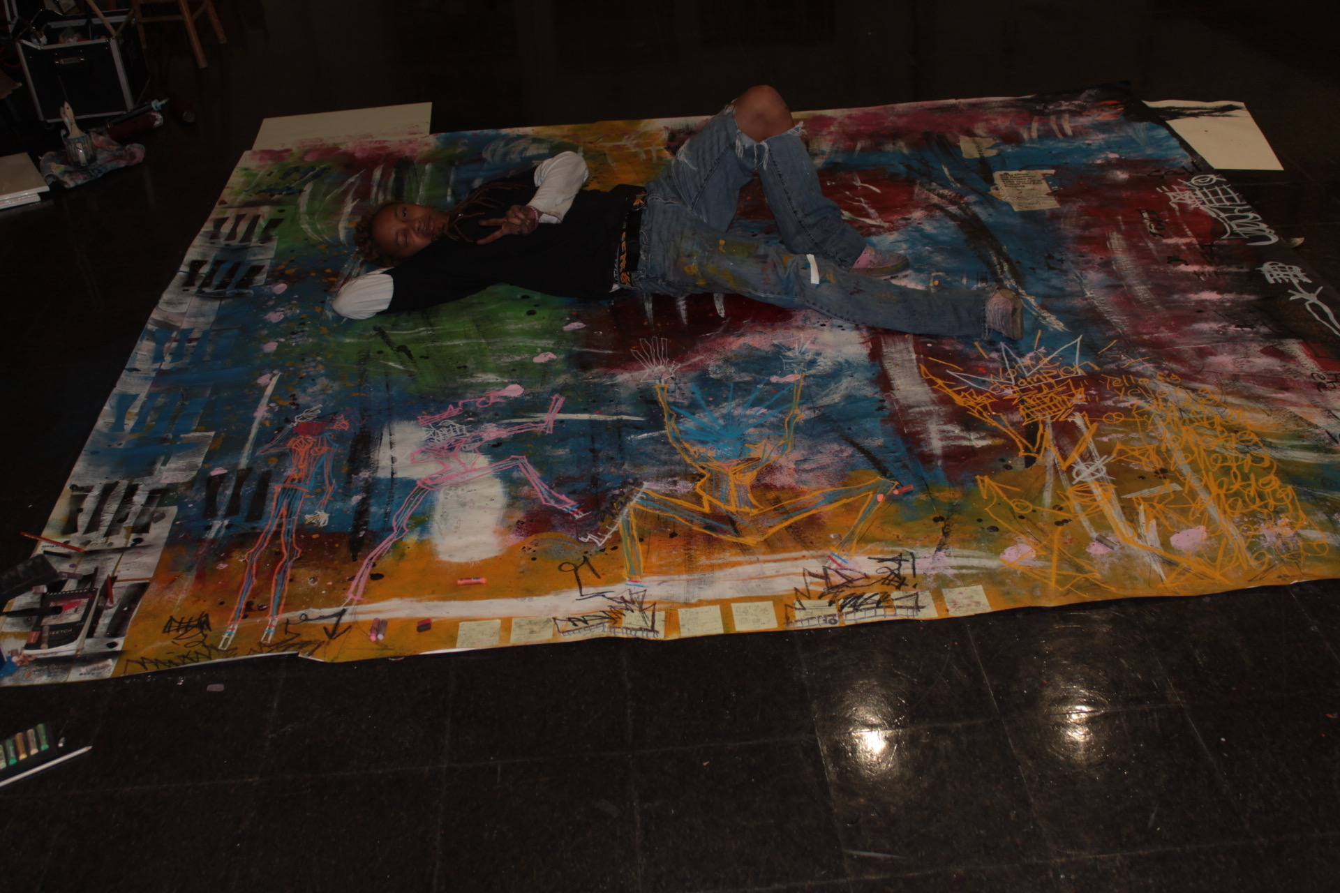 Person lying on a large, colorful puzzle on a dark floor, with a white object nearby, in a dimly lit room.