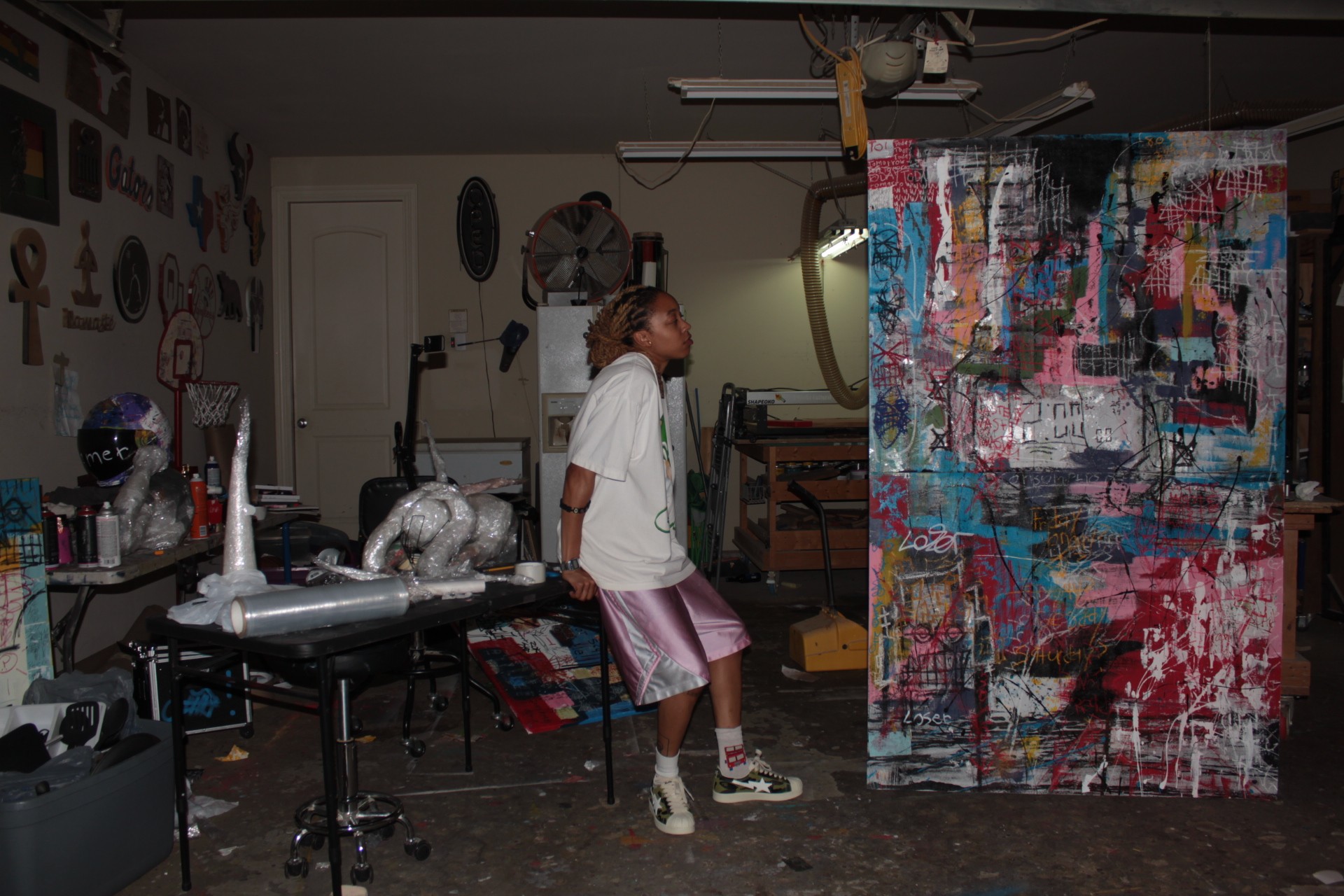 Person sitting on a stool in a cluttered room with artwork and supplies, facing a colorful abstract painting.
