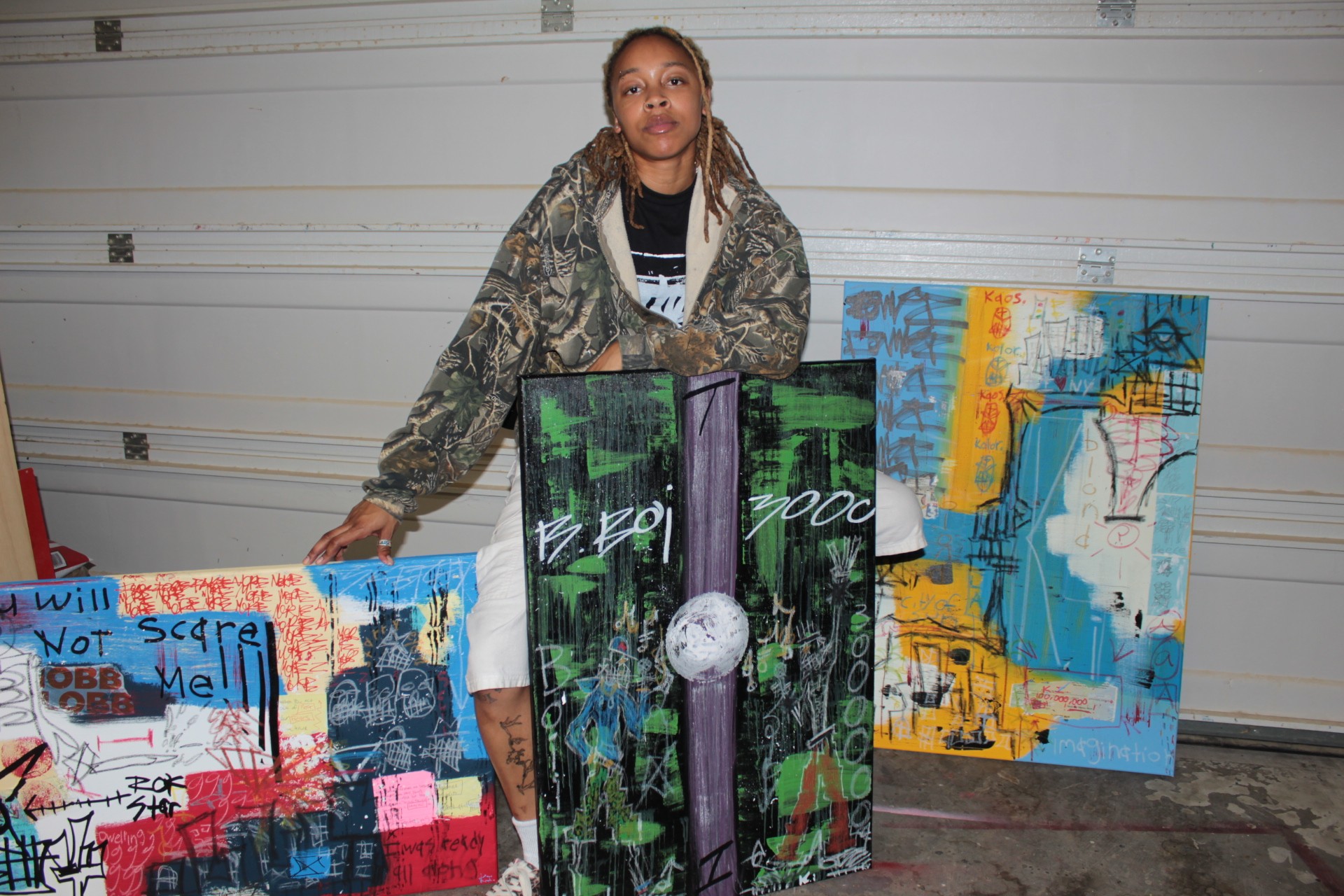 Person standing behind three colorful abstract paintings in a garage, smiling, with a white garage door in the background.