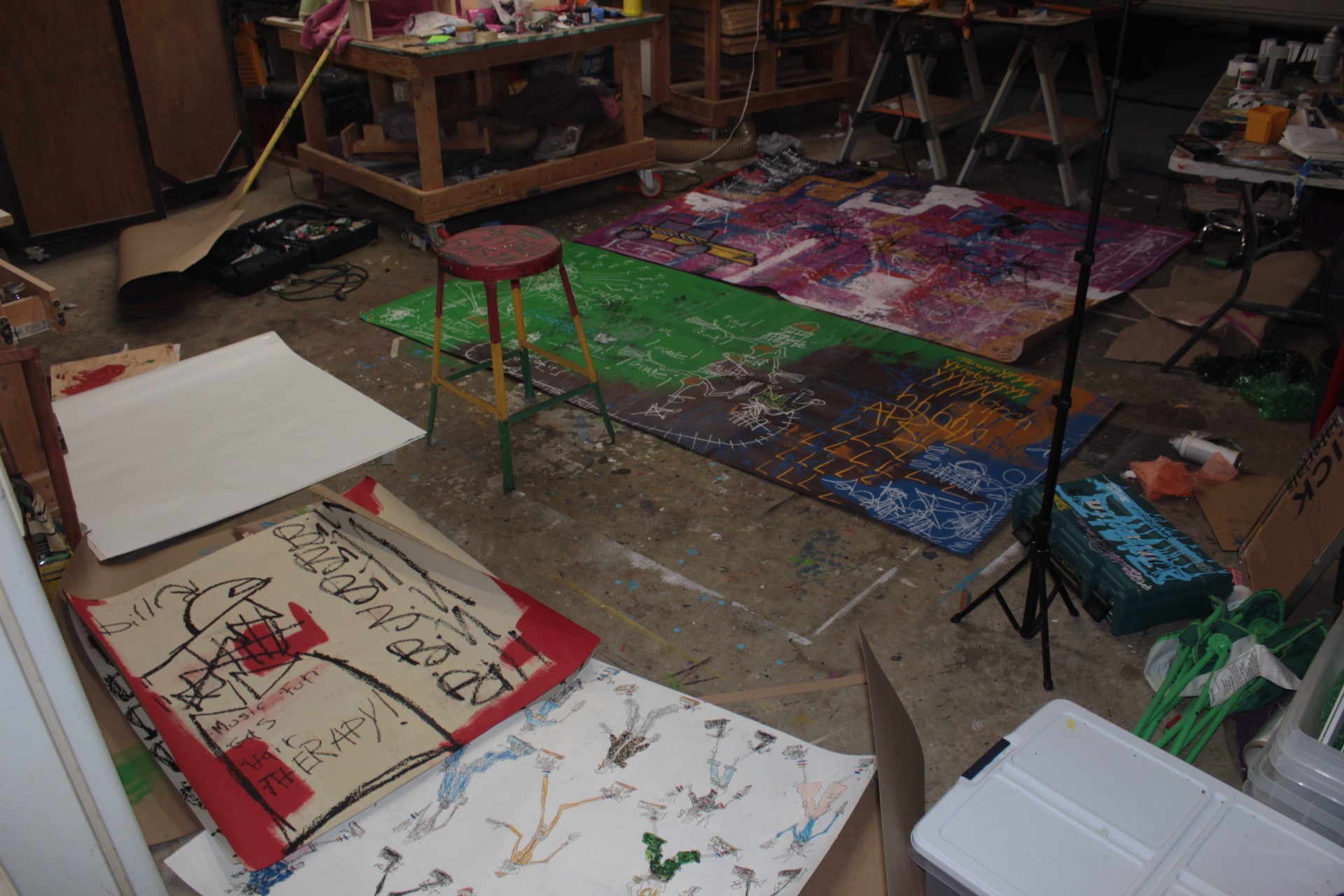 Art supplies and colorful paintings in a cluttered studio with rugs and furniture.