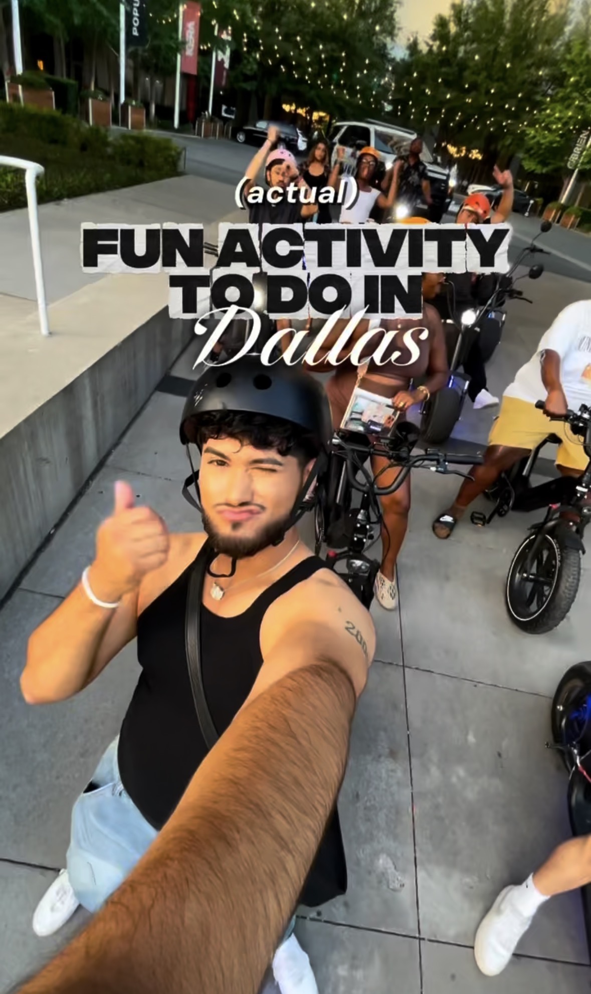 Young man taking a selfie outdoors with bicycles and people in the background, smiling and giving a thumbs-up gesture.