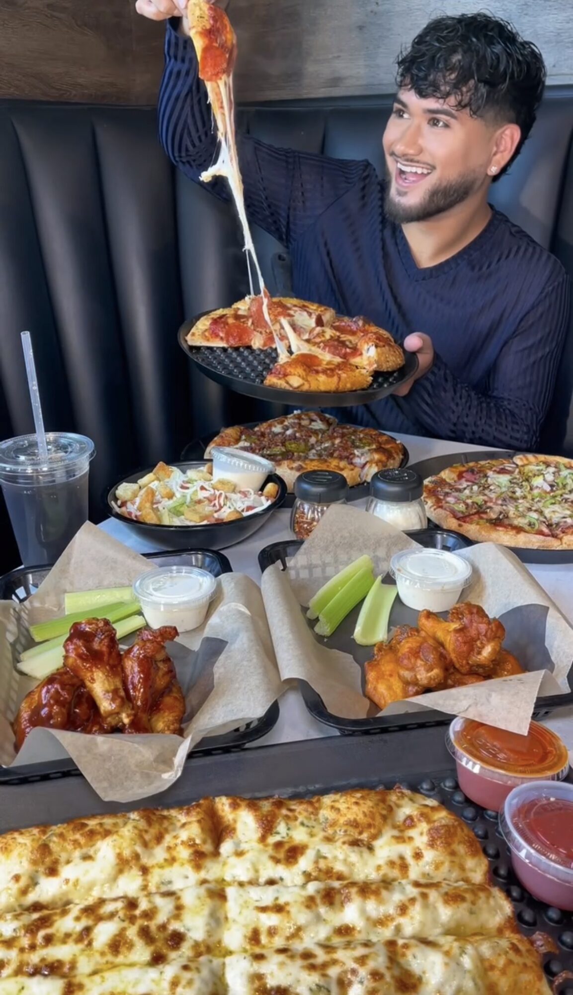 Man smiling at table with pizza, chicken wings, and drinks, holding a pizza slice with cheese stretching, in a restaurant setting.