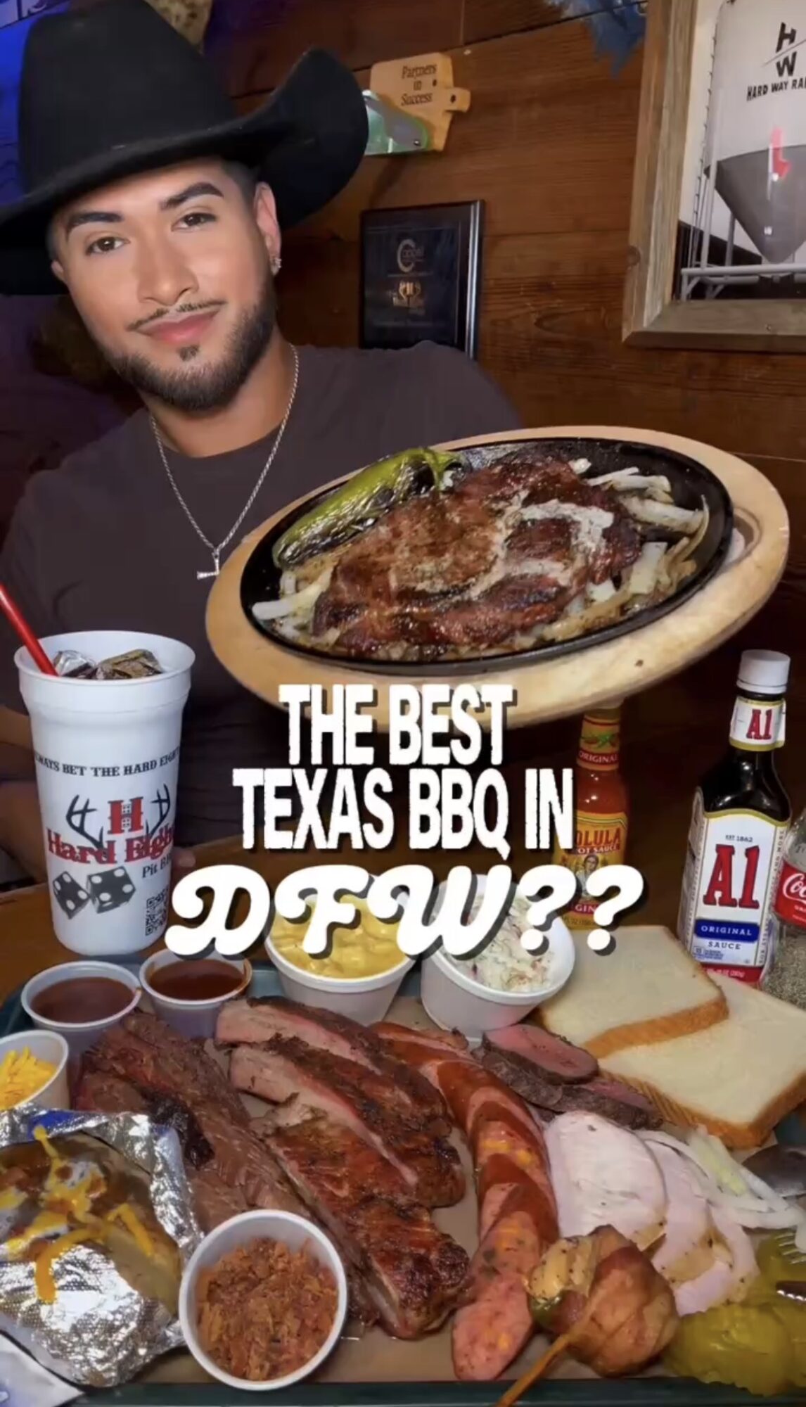 Man with a hat and beard smiling at a table with barbecue, sauces, and drinks, in a restaurant setting.