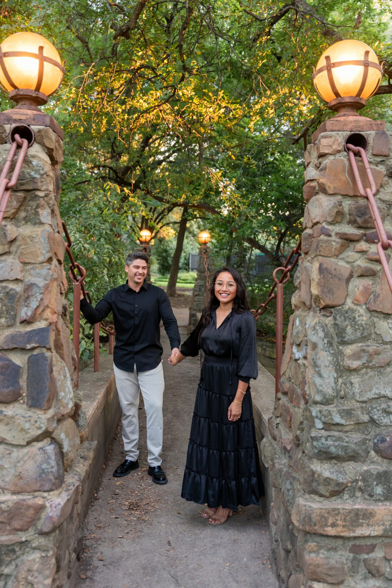 A man and woman holding hands on a stone bridge with lanterns, trees, and greenery in the background.